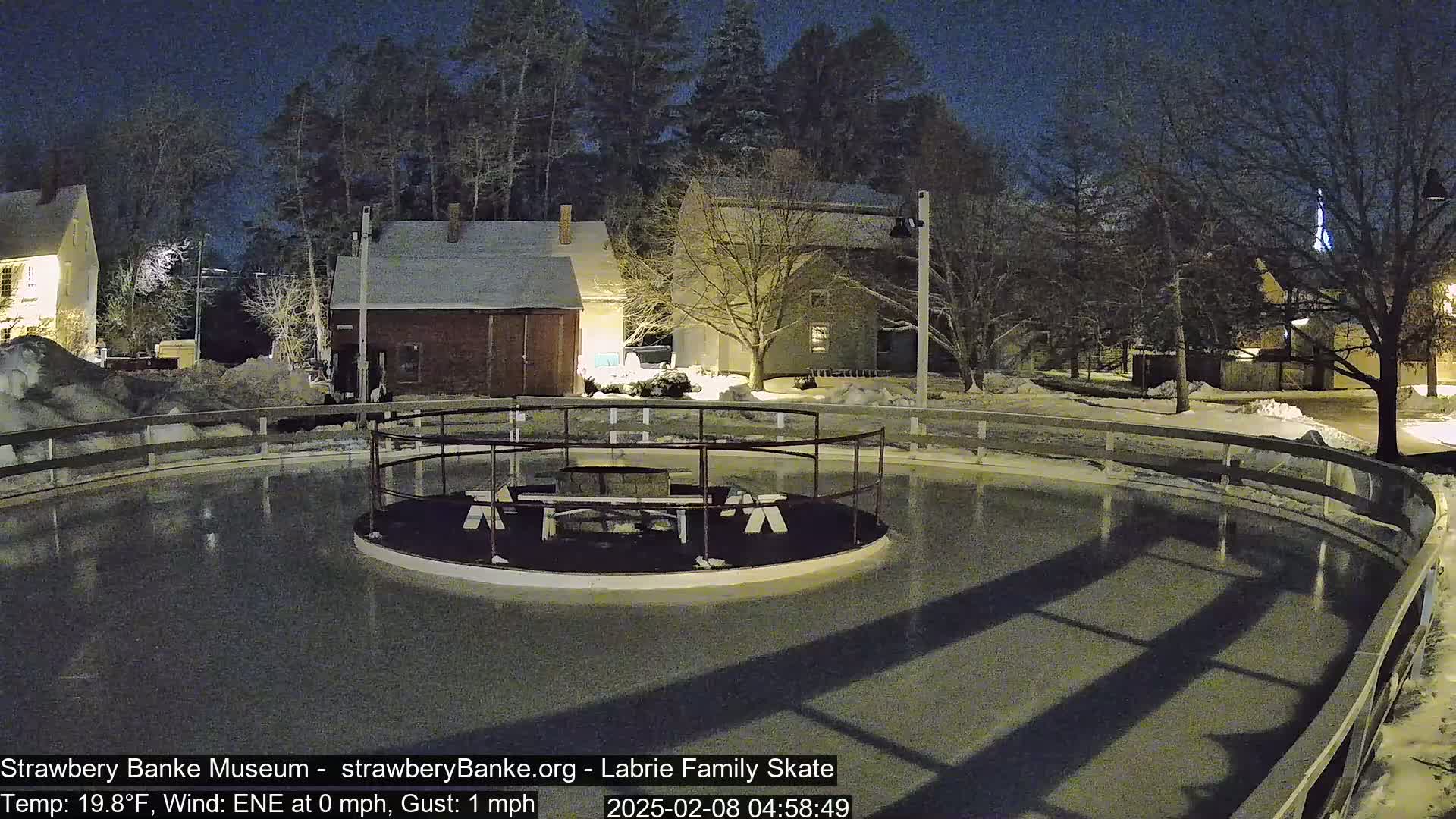 An outdoor ice rink surrounded by a low fence and lit buildings at night in snowy conditions.