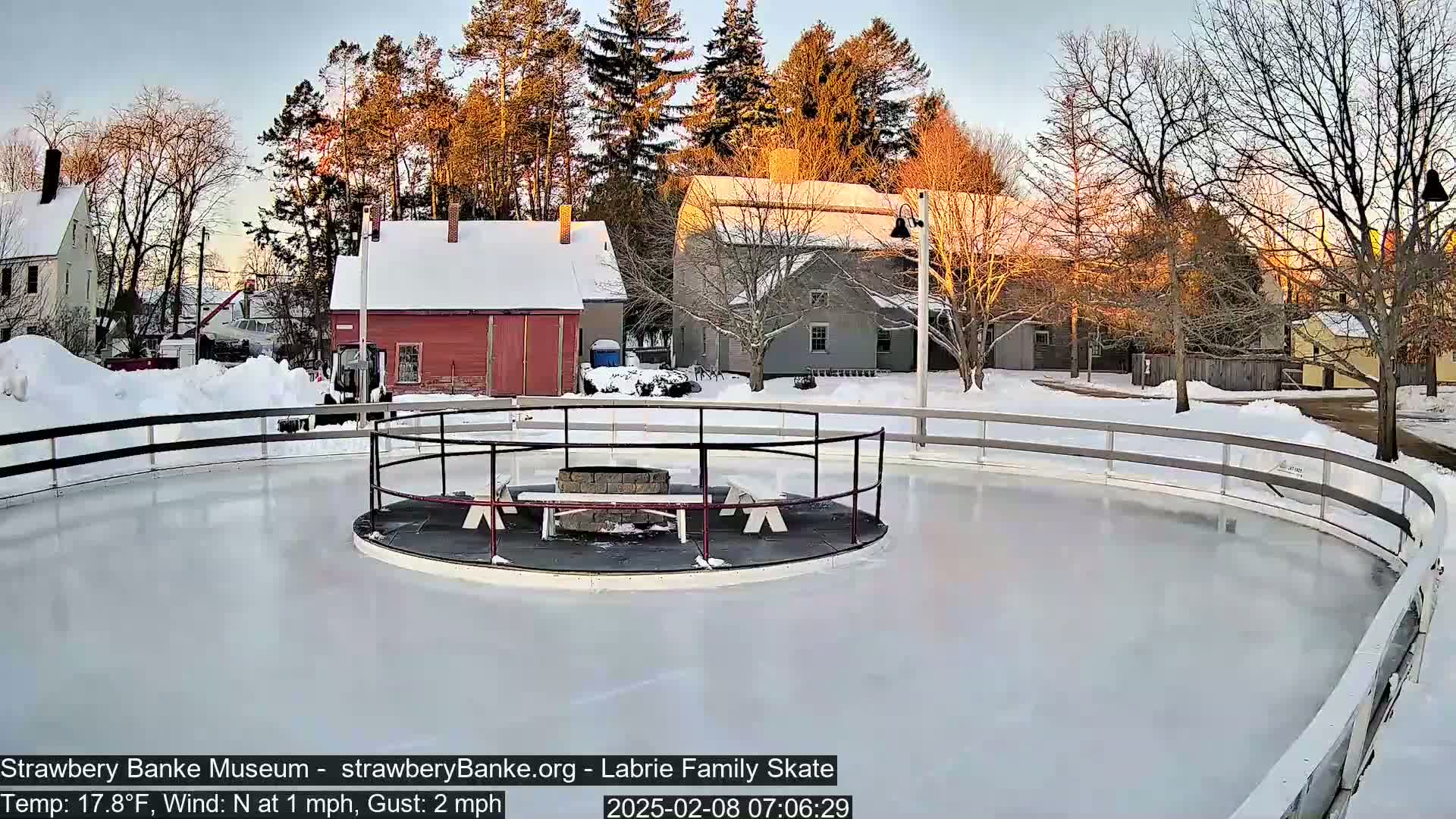A snow-covered outdoor ice skating rink is surrounded by snow-covered buildings and trees at sunset.