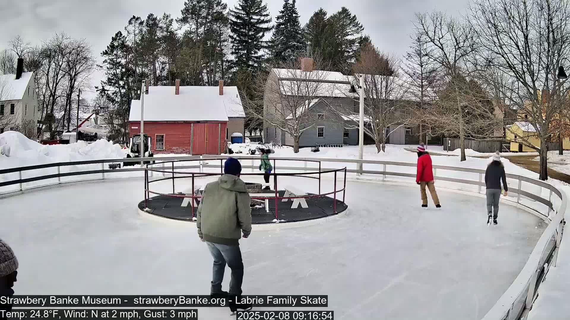 Several people ice skate on an outdoor rink surrounded by snow-covered buildings and trees on a cold, windy day.