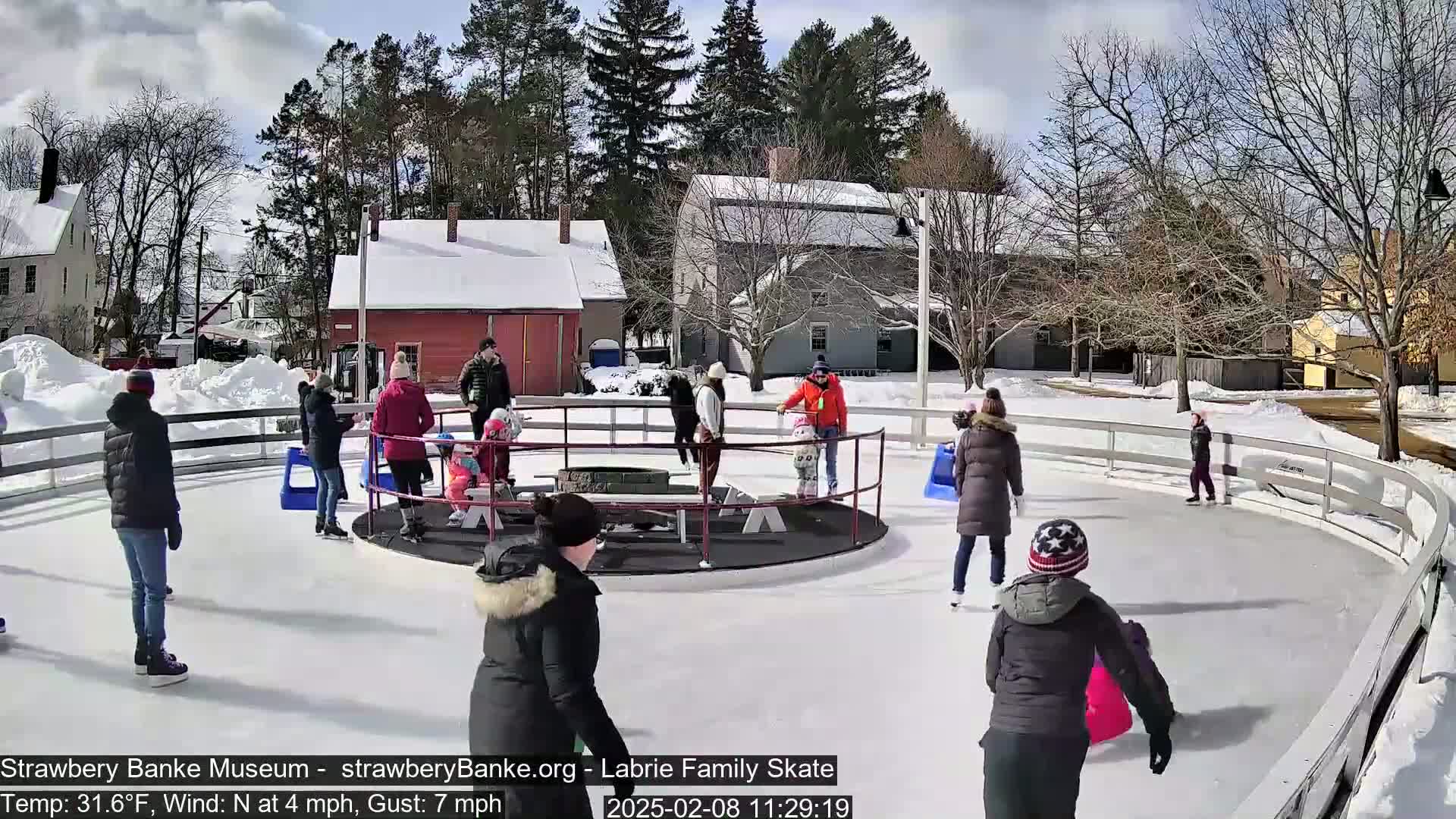 People ice skate on an outdoor rink on a sunny, cold day.