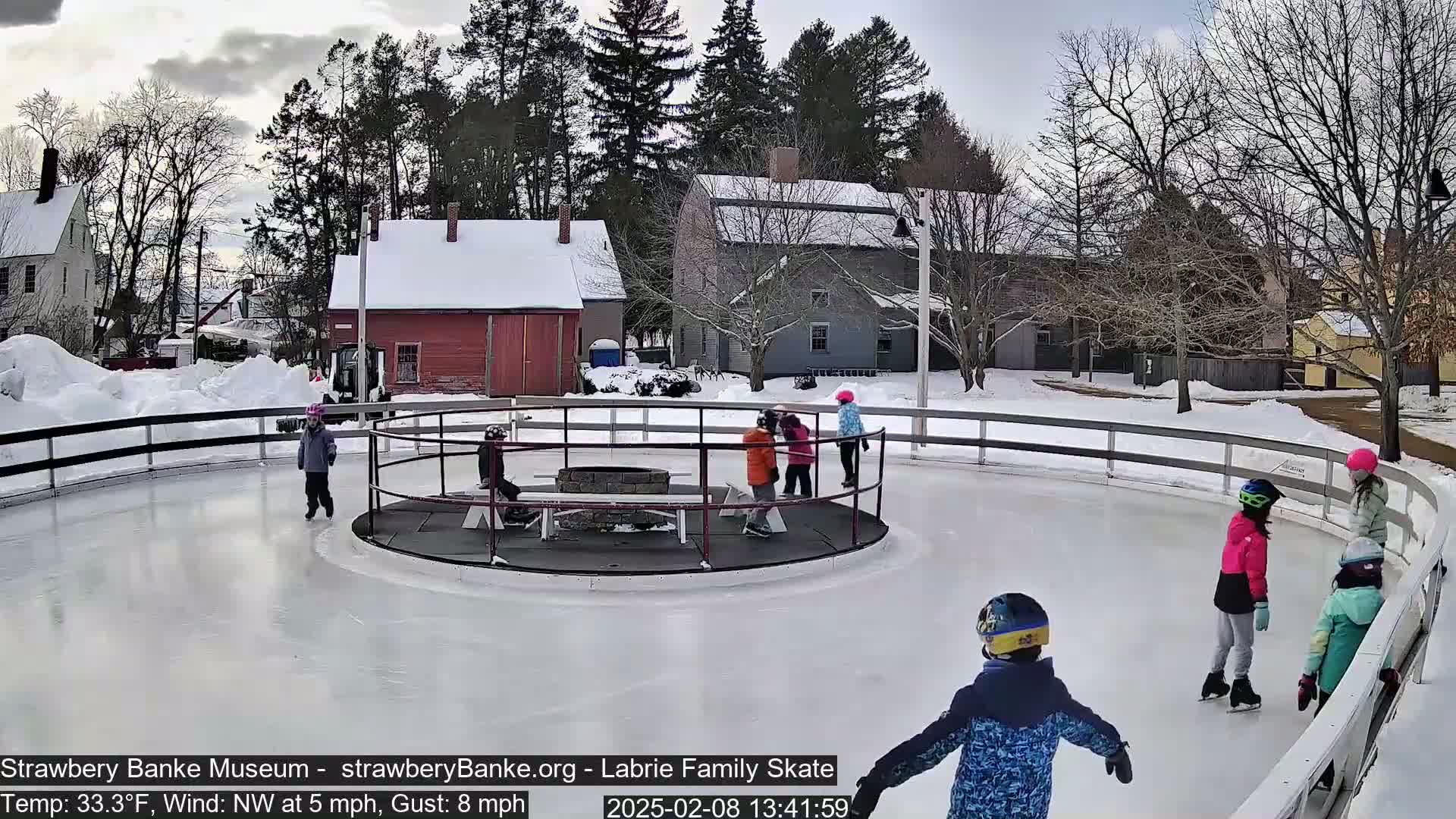 Children ice skate on an outdoor rink surrounded by snow-covered buildings and trees in a cold, windy setting.