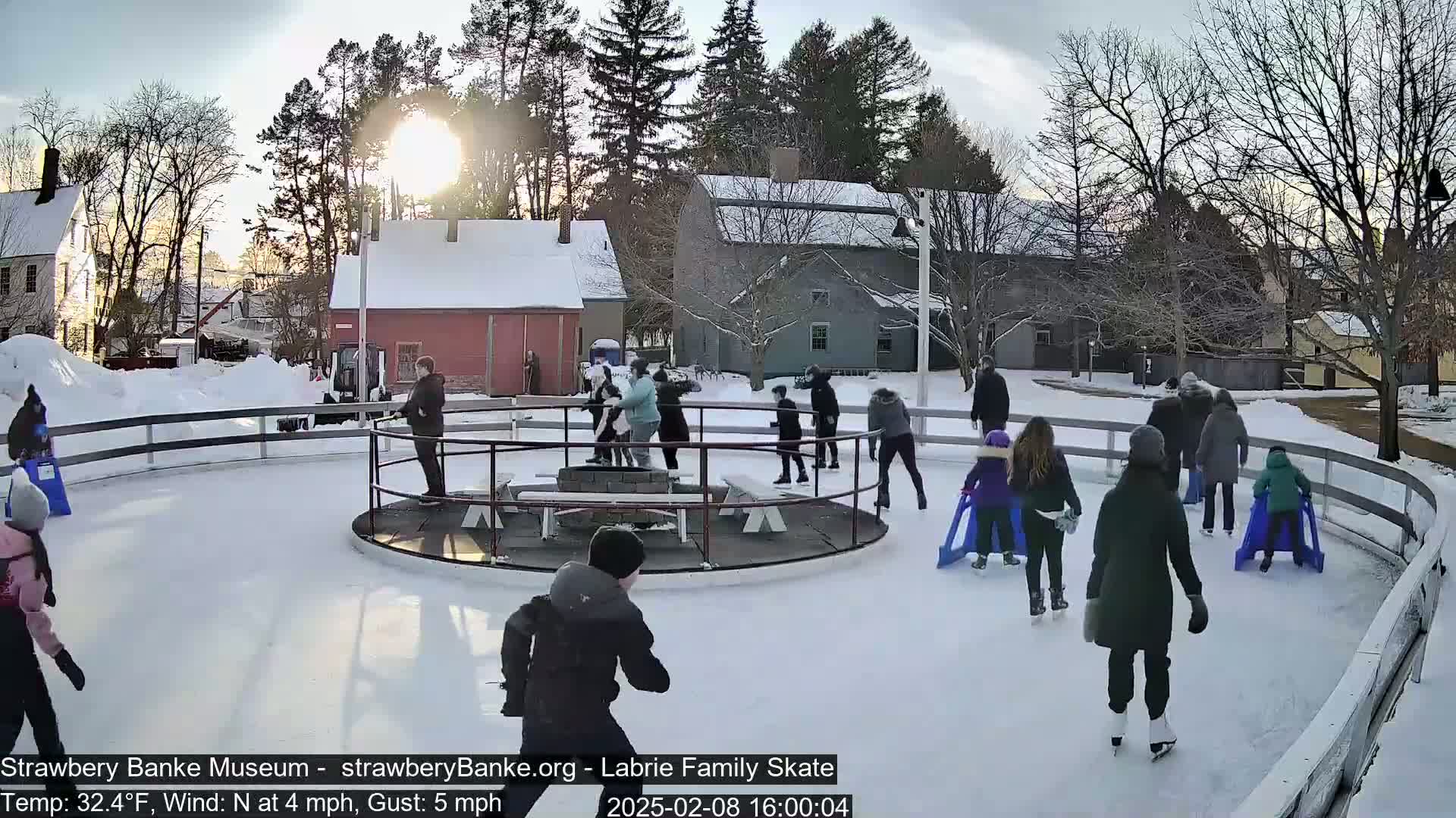 Many people ice skate on an outdoor rink surrounded by snow-covered buildings and trees under a partly sunny sky.