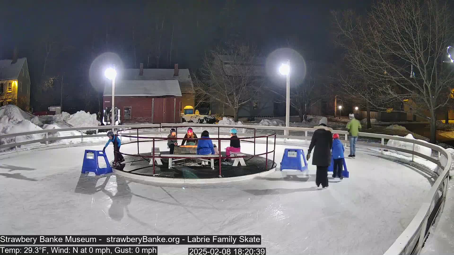 Several people ice skate on an outdoor rink at night under bright lights, with snow on the ground and calm weather.