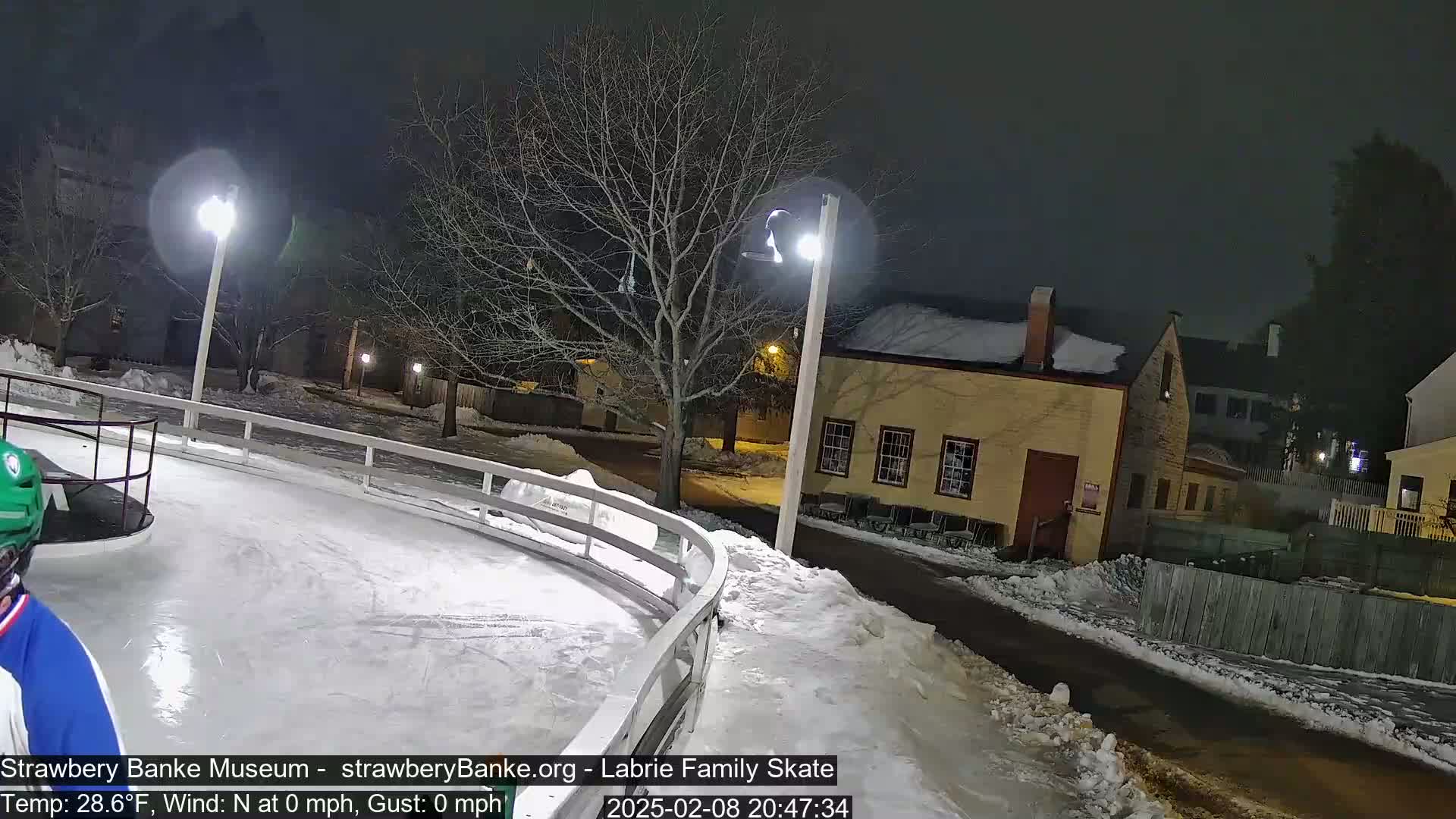 A person ice skates at night on an outdoor rink near a snow-covered street and buildings under bright lights.