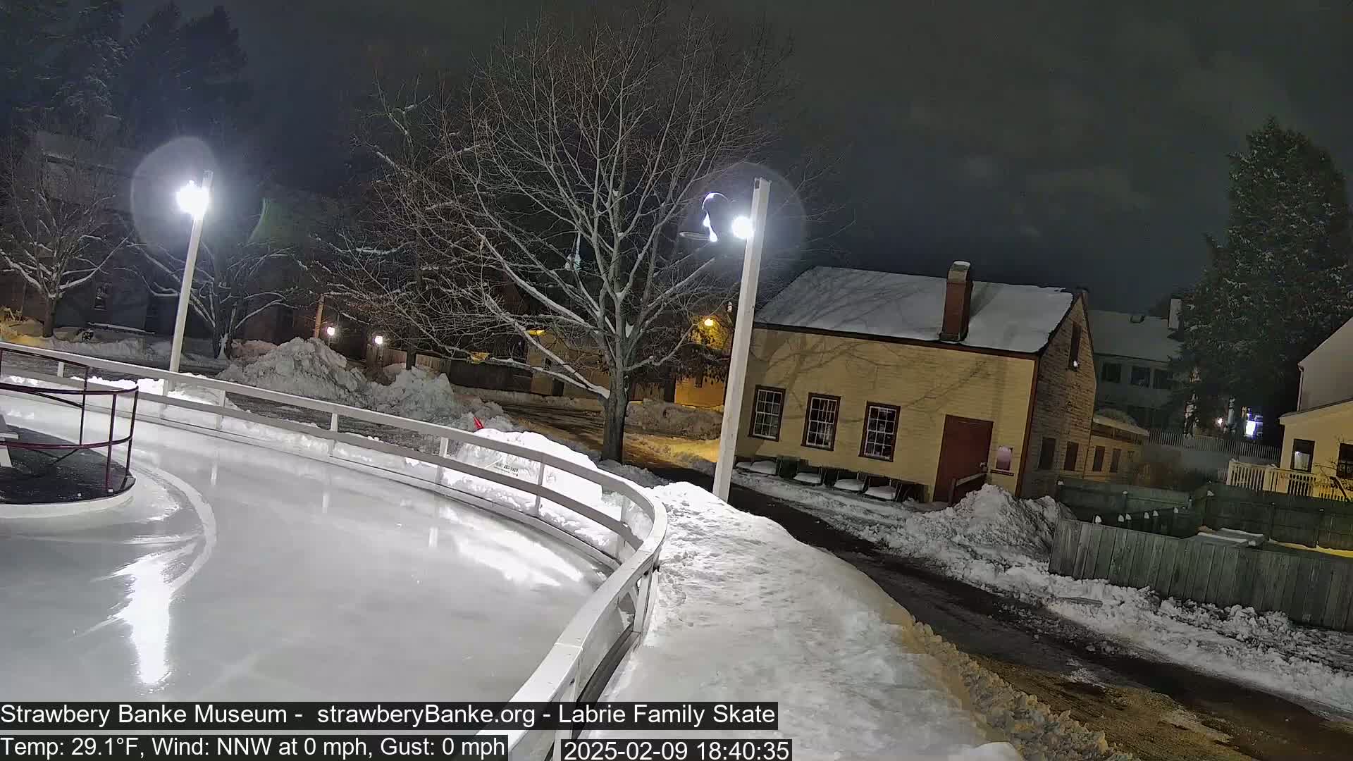 A nighttime view of a snow-covered outdoor ice rink, illuminated by lights, next to a small, yellow building and snow-covered trees.