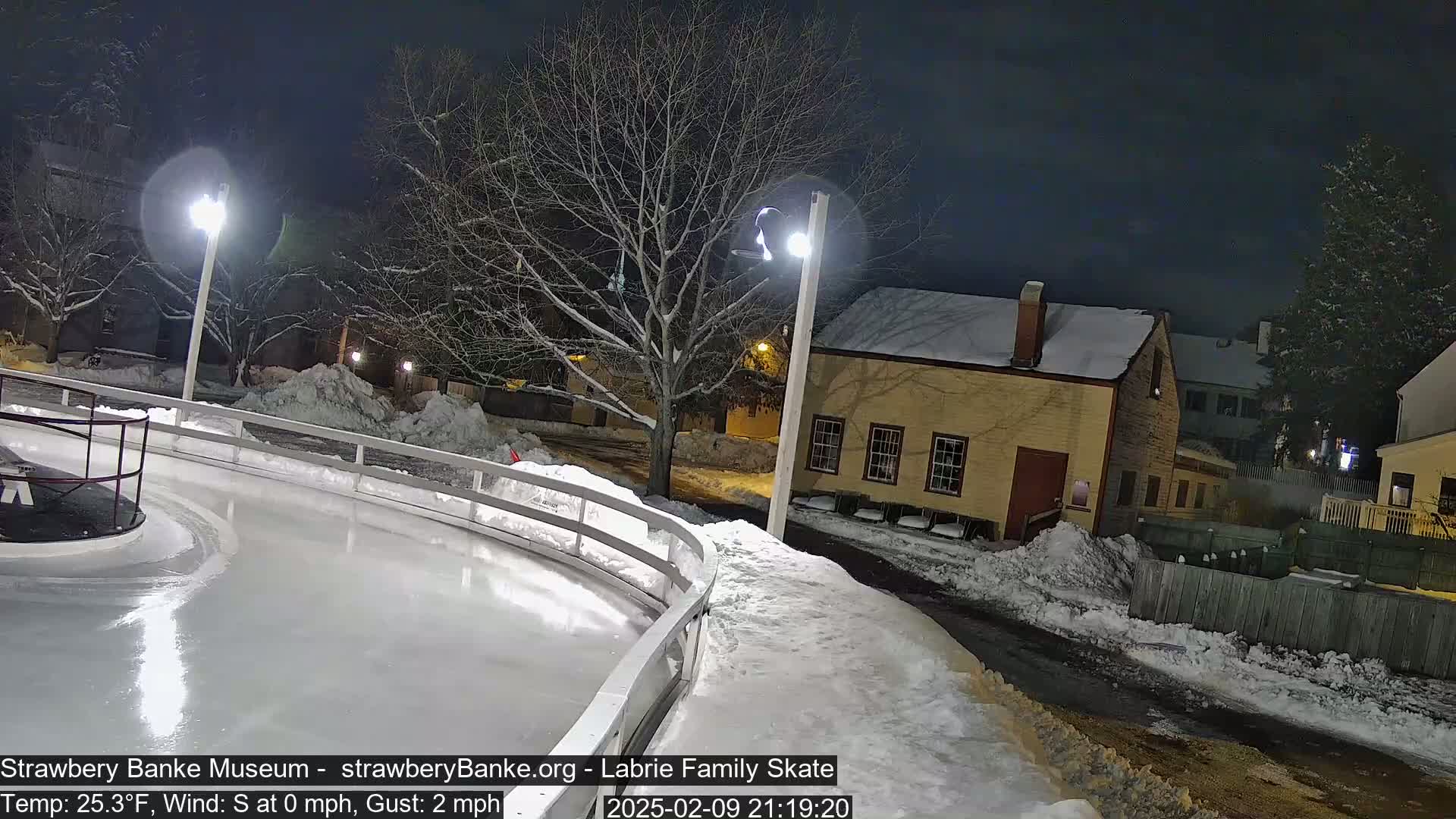 A snow-covered outdoor ice rink at night, illuminated by streetlights, is next to a yellow building and snow-covered trees.