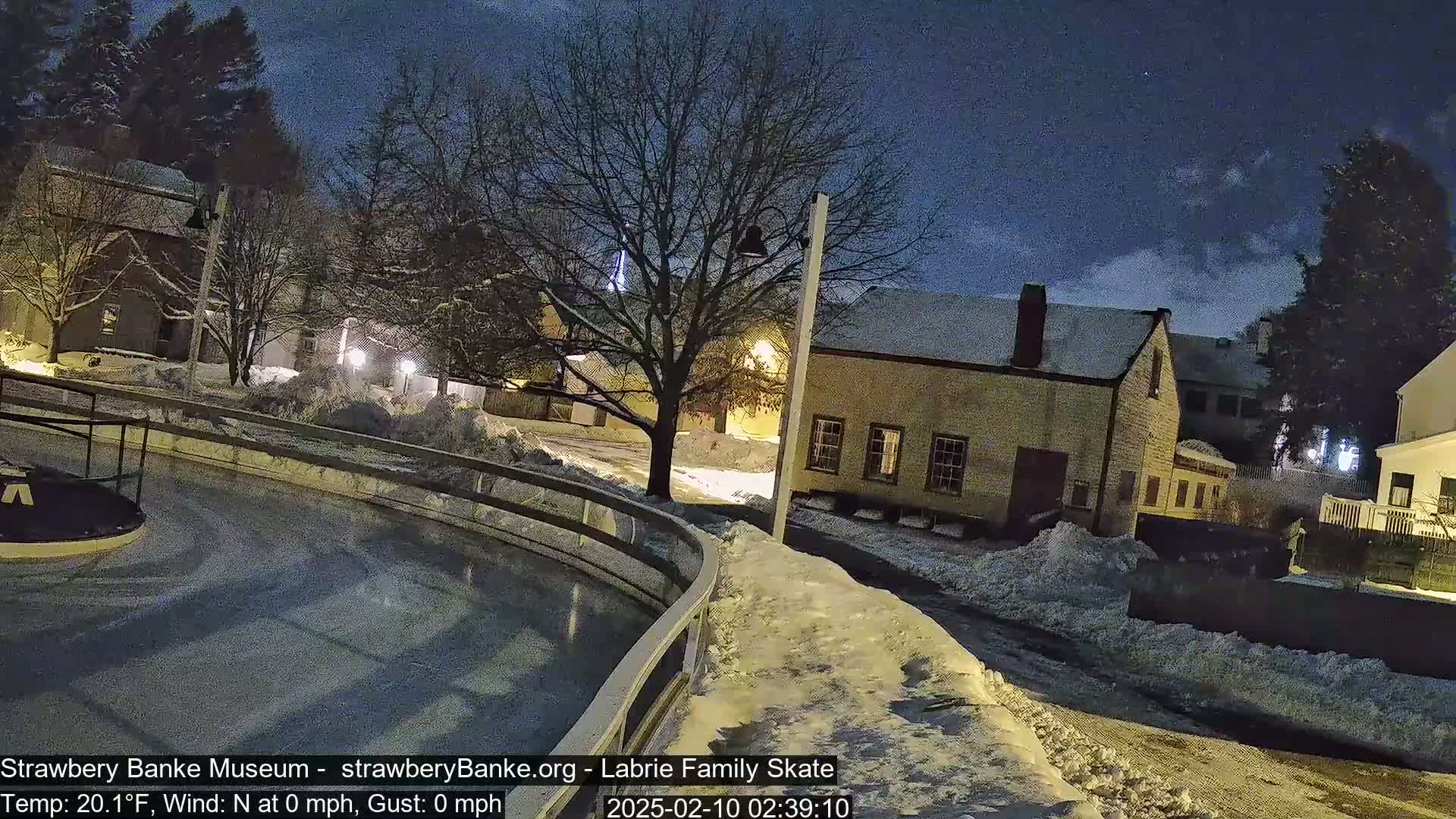 A snow-covered outdoor ice rink is next to a street with snow-covered buildings under a dark, clear night sky.
