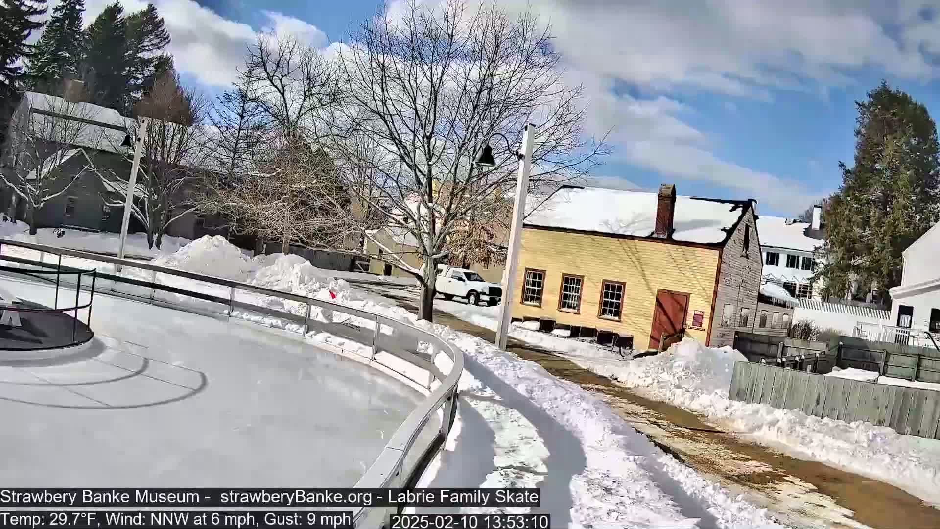 A partly sunny winter day shows a snow-covered outdoor ice rink next to a yellow building and snow-covered streets.