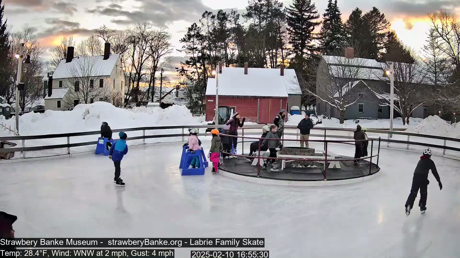 Children ice skate on an outdoor rink at sunset, surrounded by snow-covered buildings and trees, in a cold, windy environment.