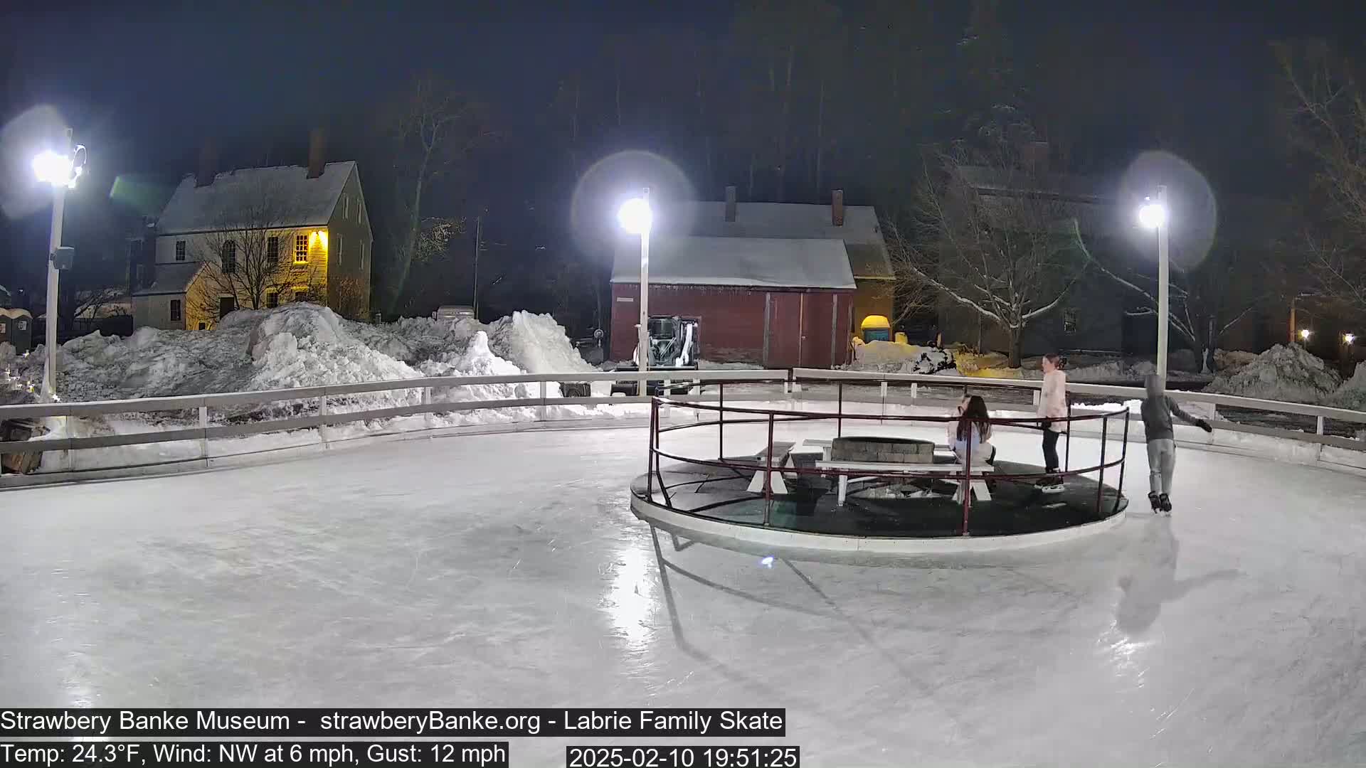 Three people ice skate at night on an outdoor rink illuminated by lights, surrounded by snow-covered buildings and piles of snow in a cold, windy environment.