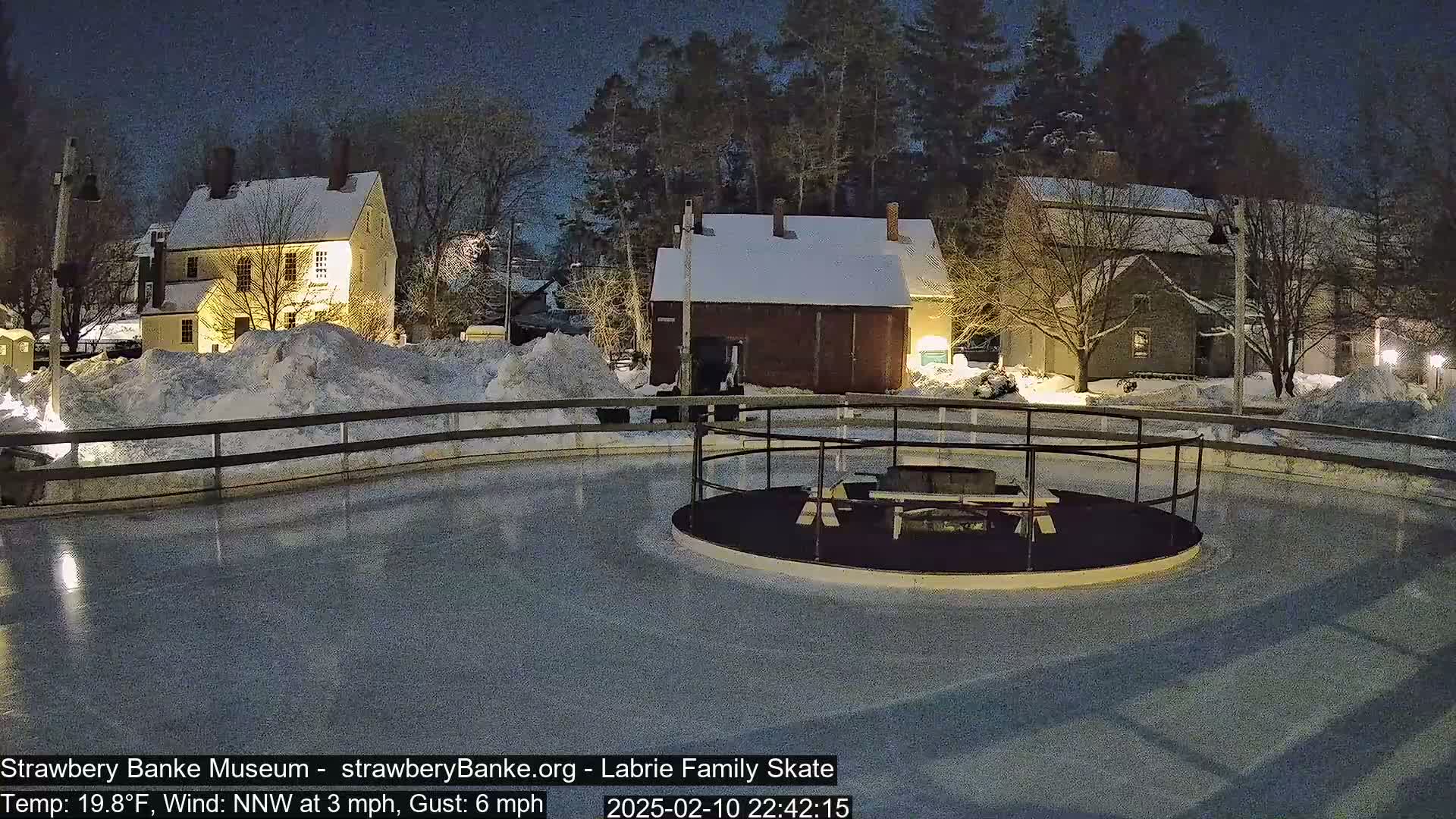 A nighttime view of a snow-covered outdoor ice skating rink, illuminated by lights, with several historic-looking buildings in the background under a dark sky.
