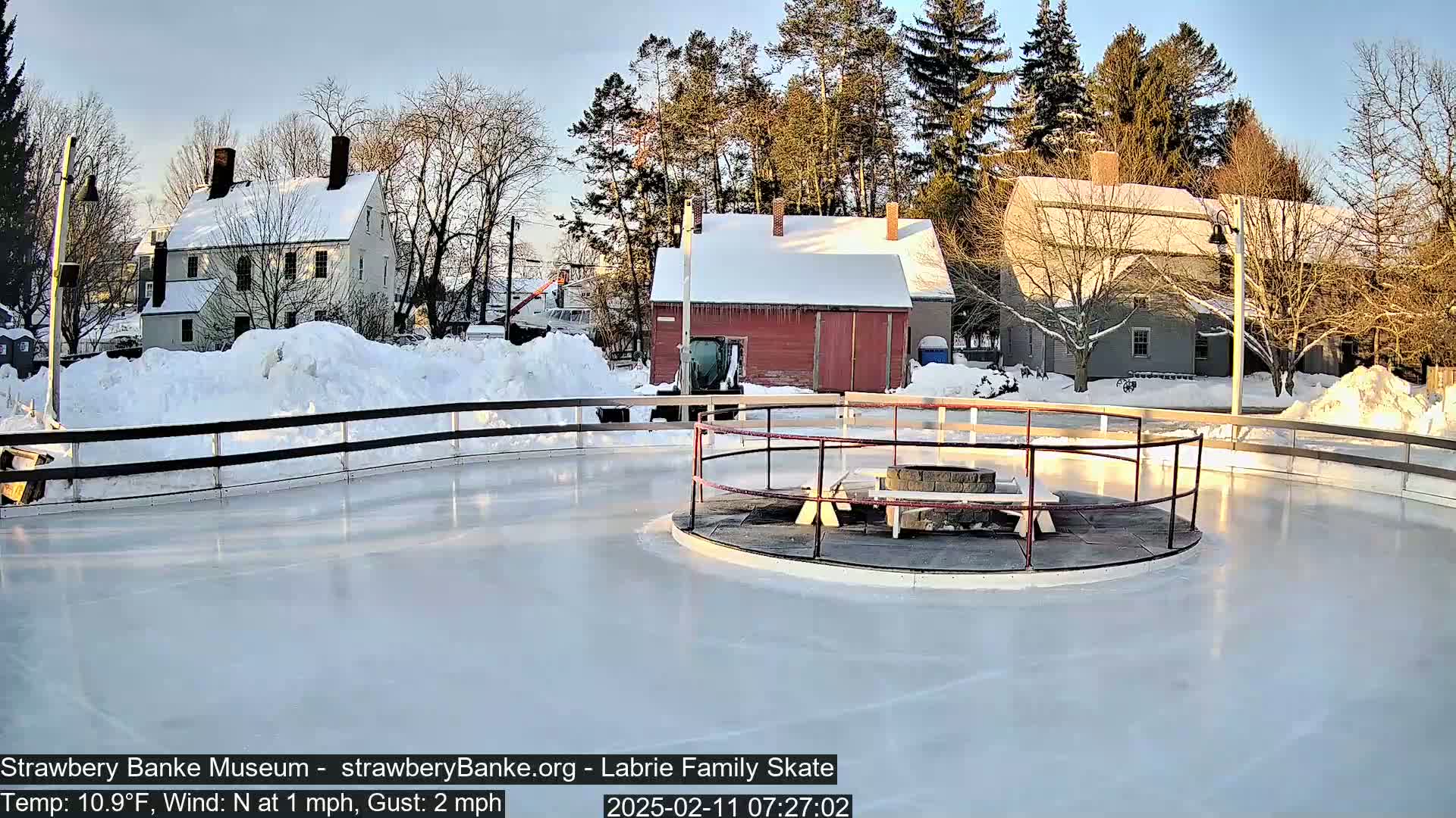 A snow-covered outdoor ice skating rink with a fire pit in the center, surrounded by historic-looking buildings under a clear, sunny sky.