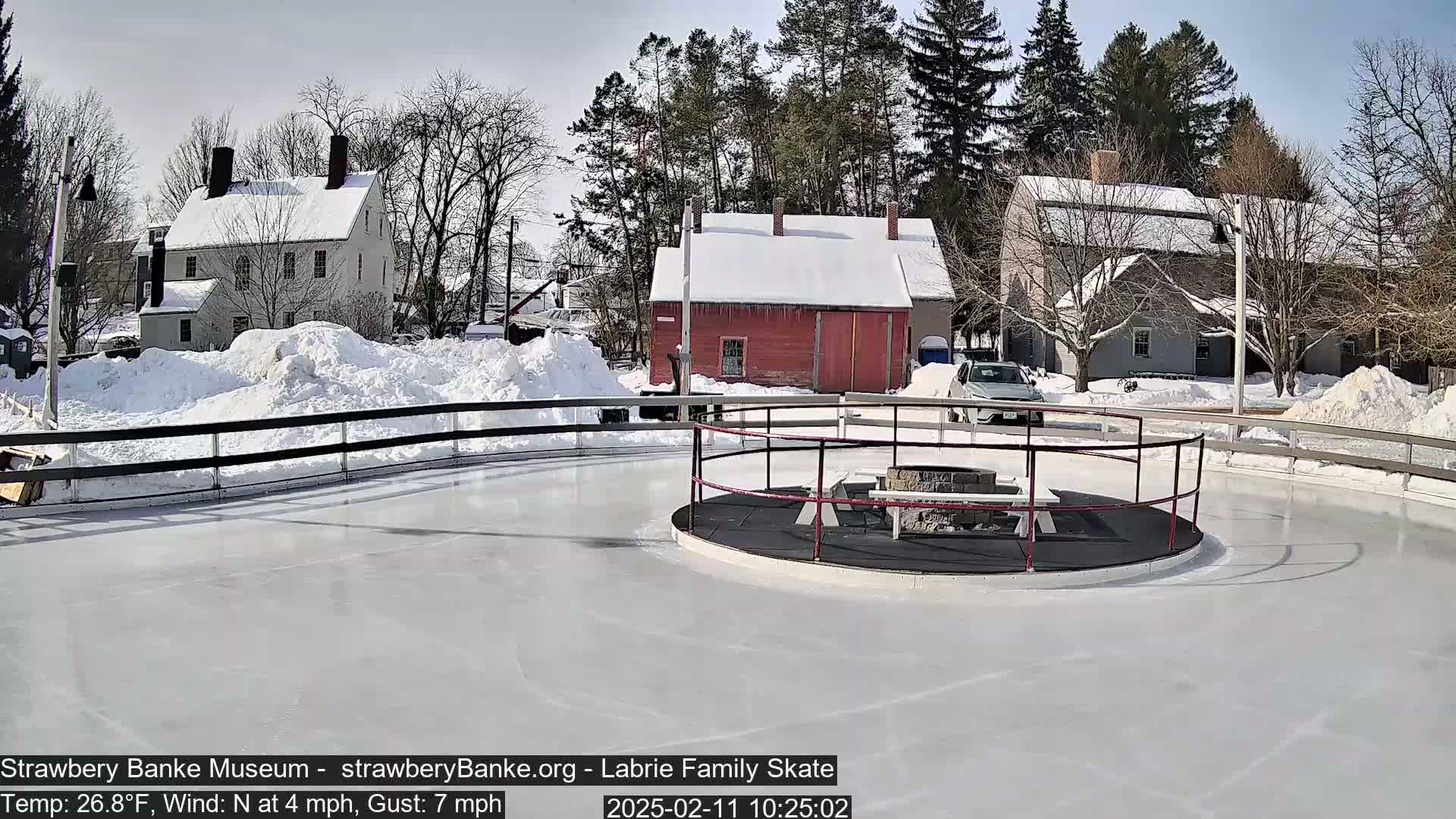 A snow-covered outdoor ice skating rink with a fire pit and surrounding buildings on a sunny day.