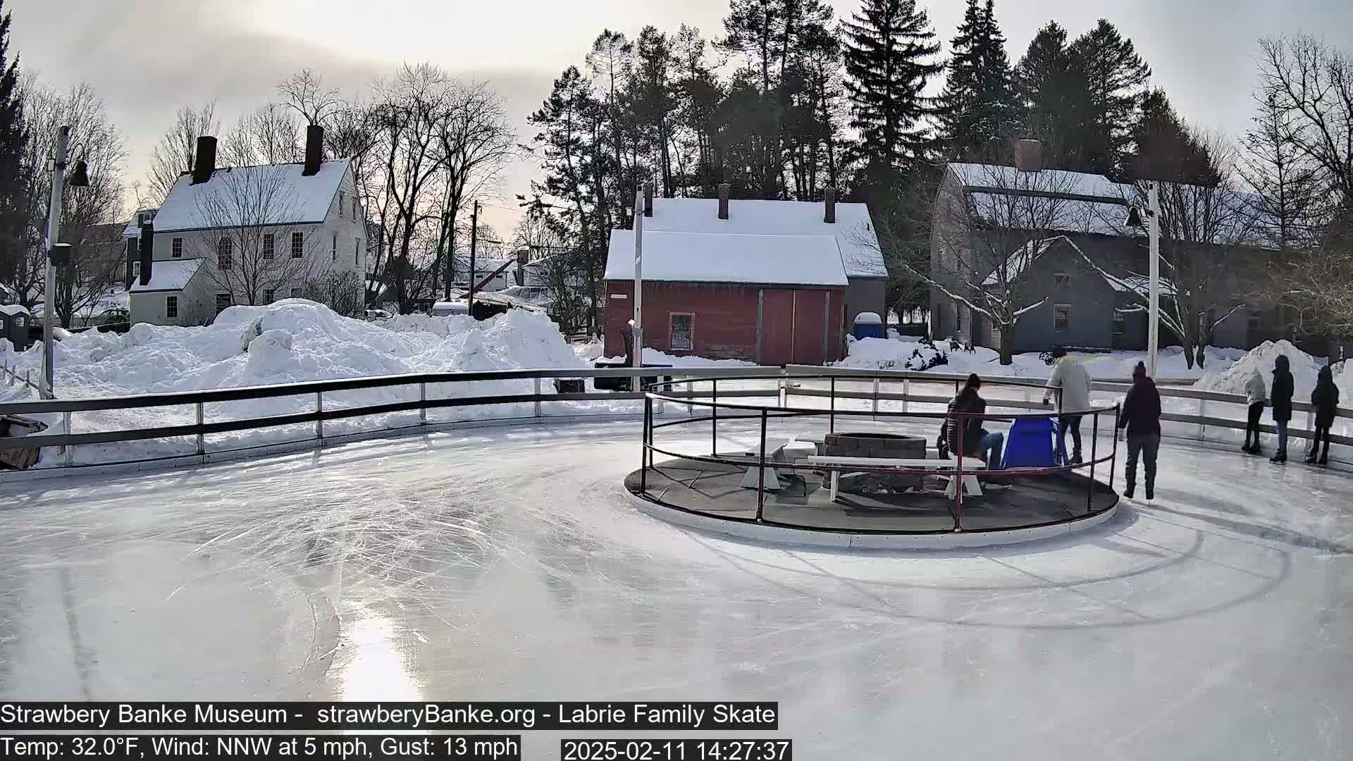 A sunny outdoor ice-skating rink, surrounded by snow-covered buildings and trees, with several people ice skating.