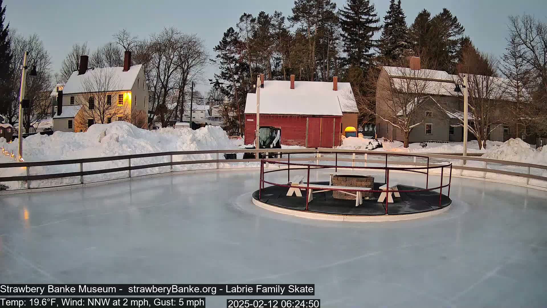 A snow-covered outdoor ice skating rink with a fire pit and benches in the center, surrounded by snow-covered buildings and trees under a twilight sky.