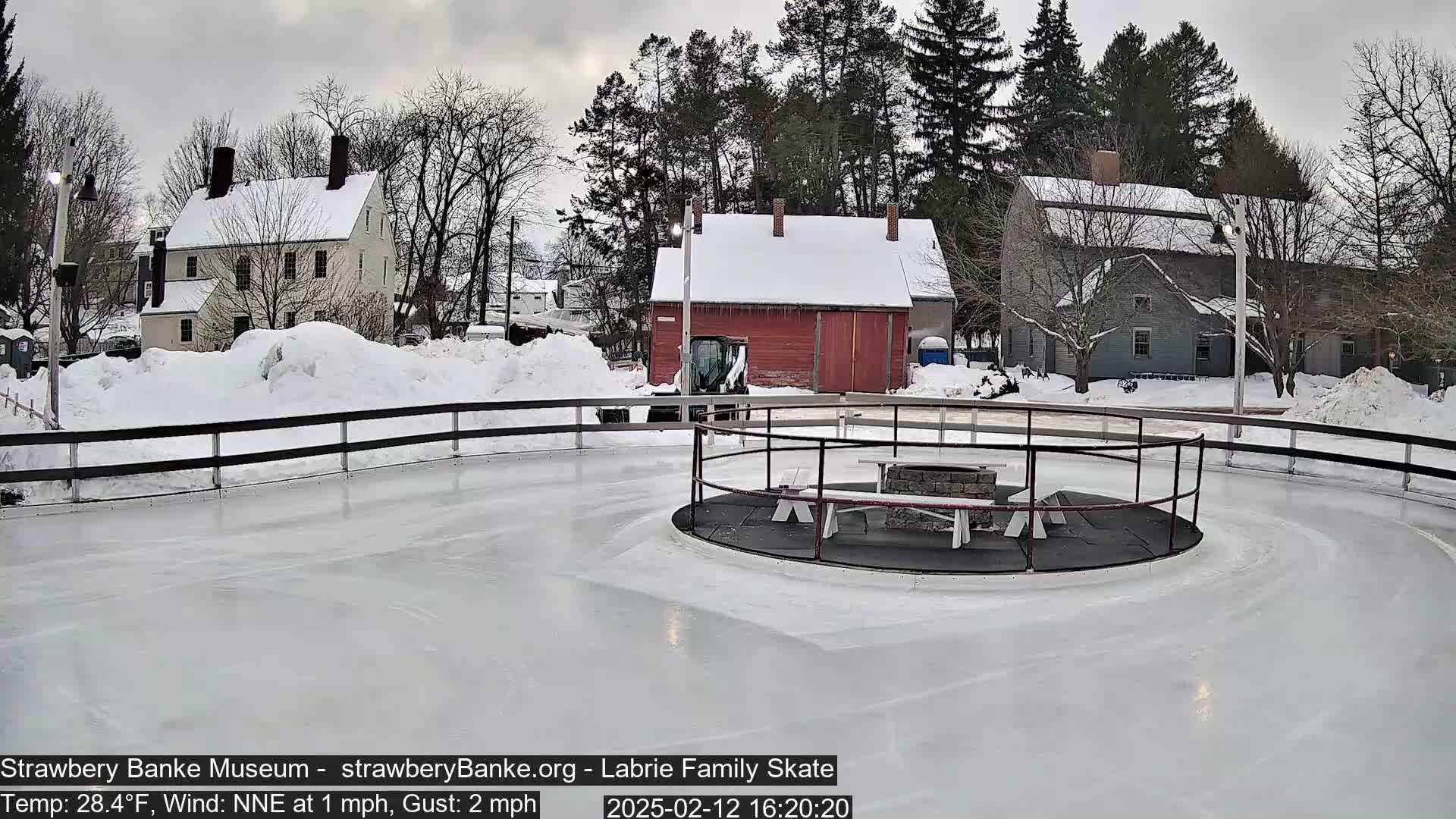 A snow-covered outdoor ice skating rink with a central fire pit and picnic tables is surrounded by buildings and trees under an overcast sky.