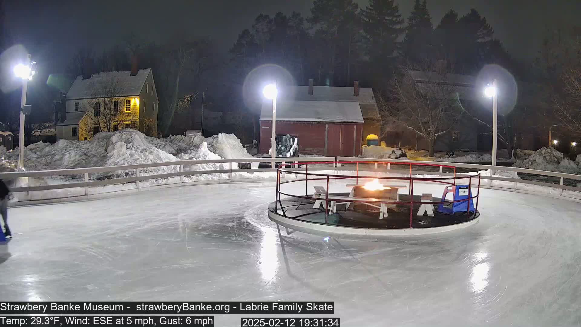 A nighttime view of an outdoor ice skating rink illuminated by lights, with a fire pit in the center, surrounded by snow and buildings.