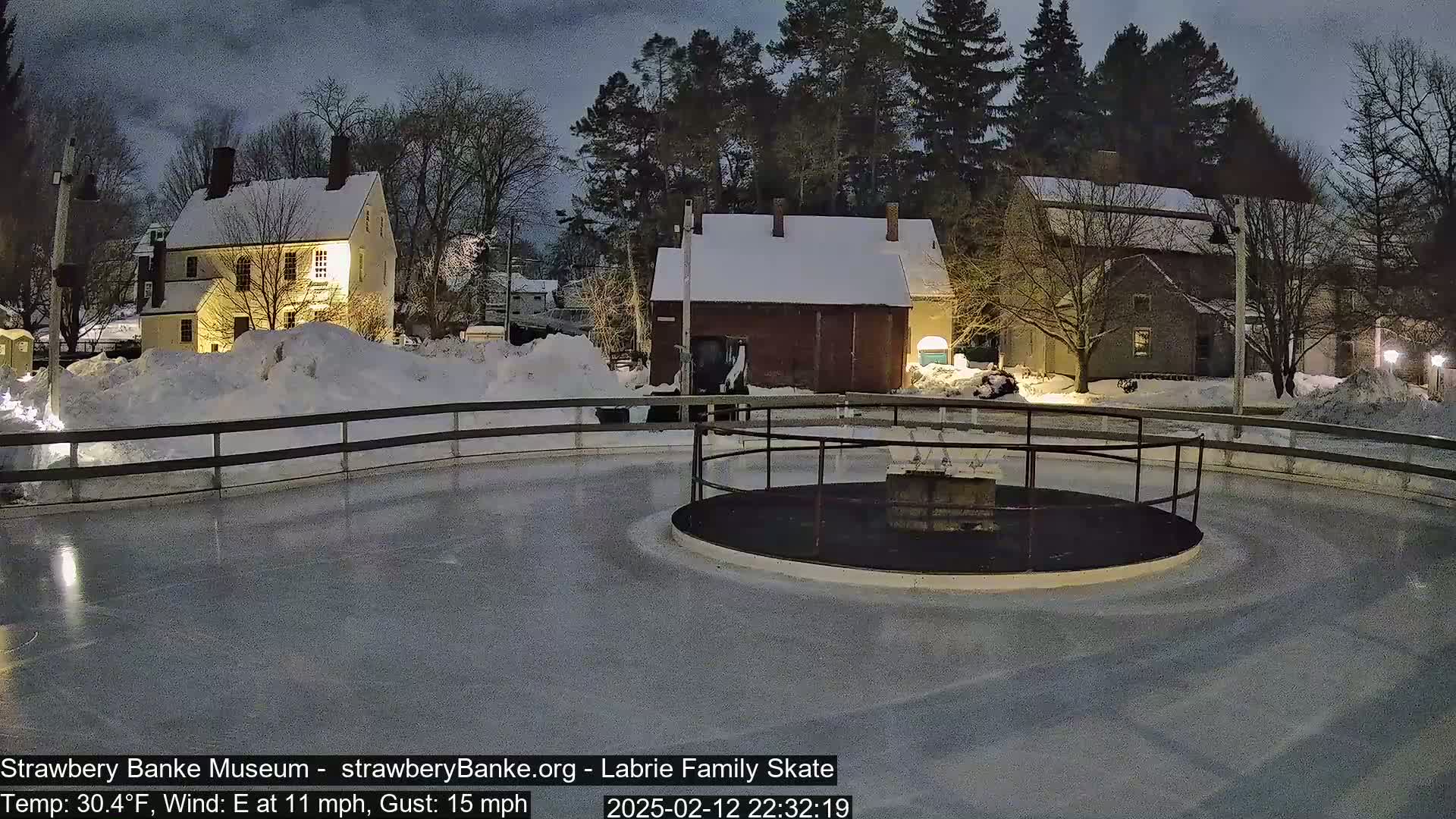A snow-covered outdoor ice skating rink at night, illuminated by lights, is surrounded by historic-looking buildings and trees.