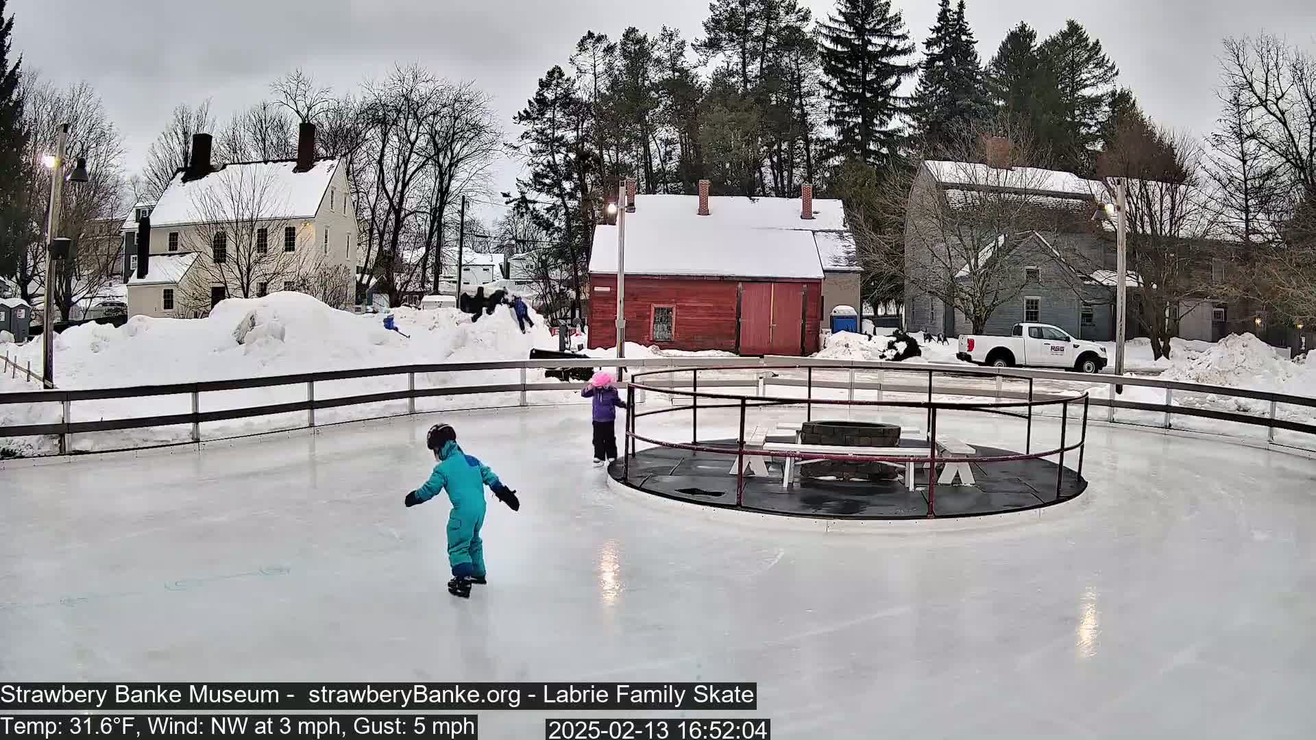 Two children ice skate on a rink surrounded by snow-covered buildings and trees under an overcast sky.