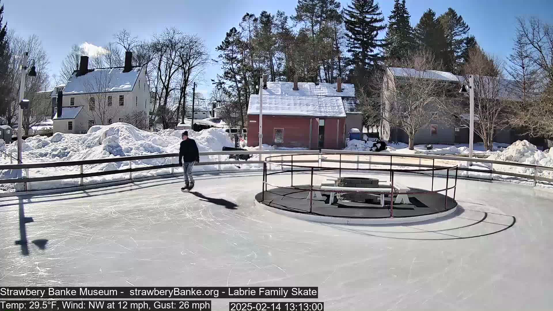 A person ice skates on a rink surrounded by snow-covered ground and buildings under a sunny sky.