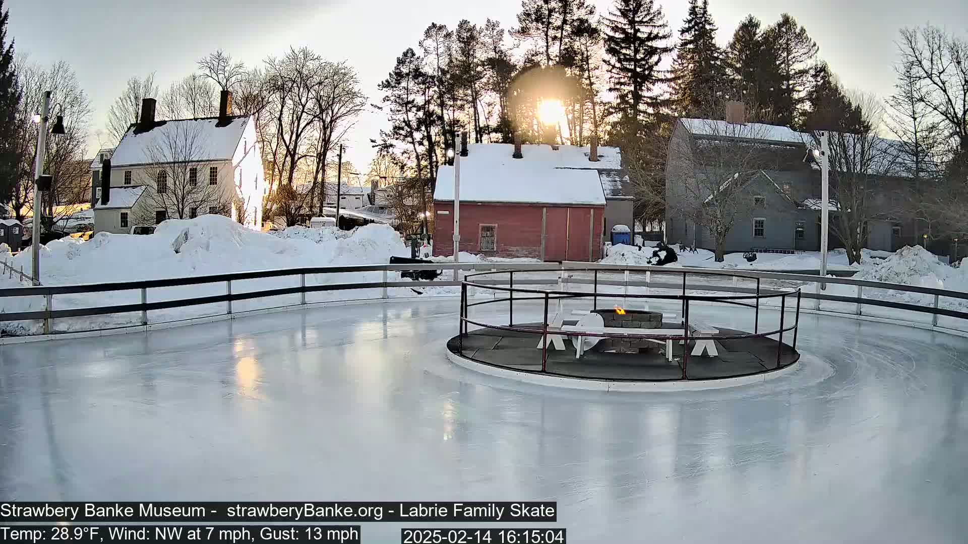 A snow-covered outdoor ice skating rink with a fire pit in the center, surrounded by historic-looking buildings under a bright, sunny sky.