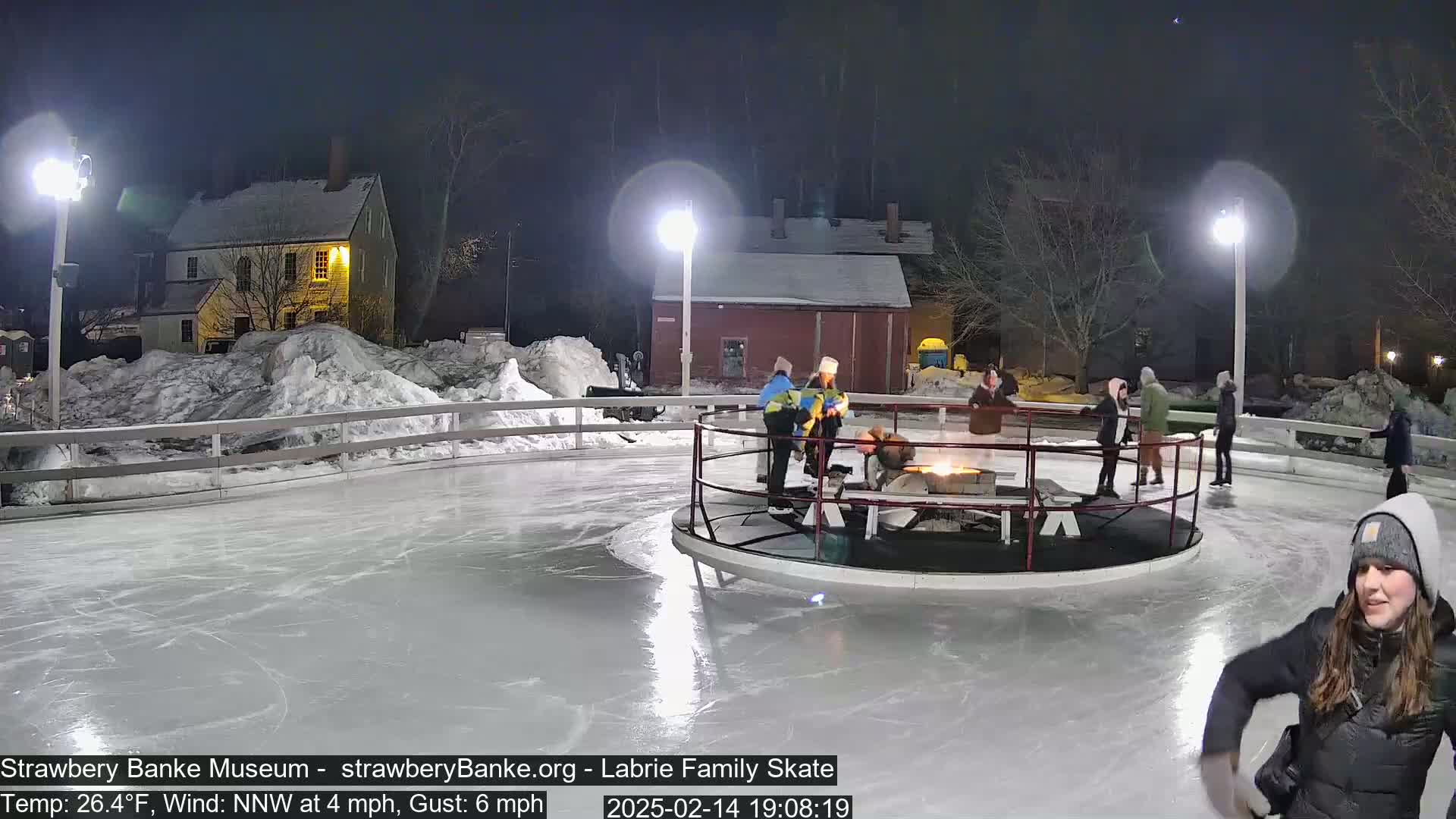 An outdoor ice skating rink at night, lit by several lamps, features several people ice skating around a central fire pit, under a clear, dark sky with a slight wind.