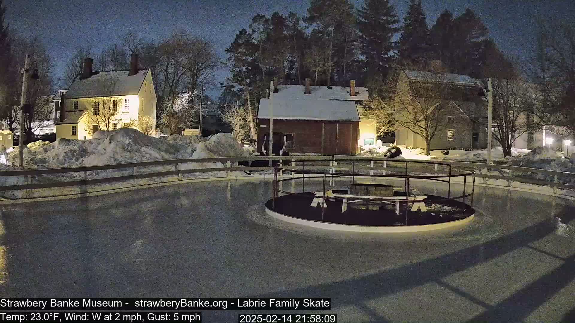 A nighttime view of a snow-covered outdoor ice rink illuminated by ambient light, surrounded by historical-looking buildings and trees under a dark sky.
