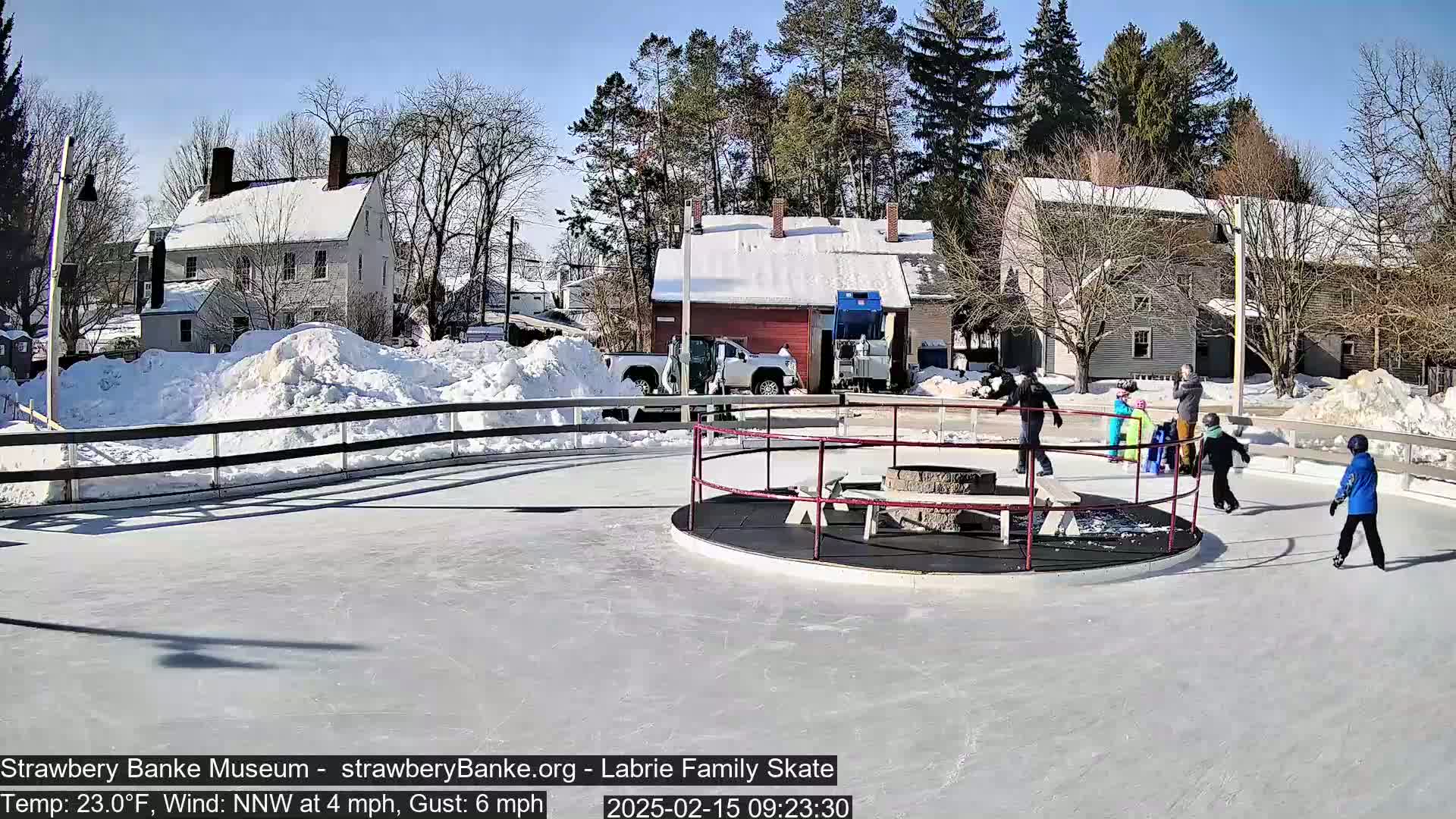 A sunny, snowy day shows several people ice skating on an outdoor rink near snow-covered buildings.
