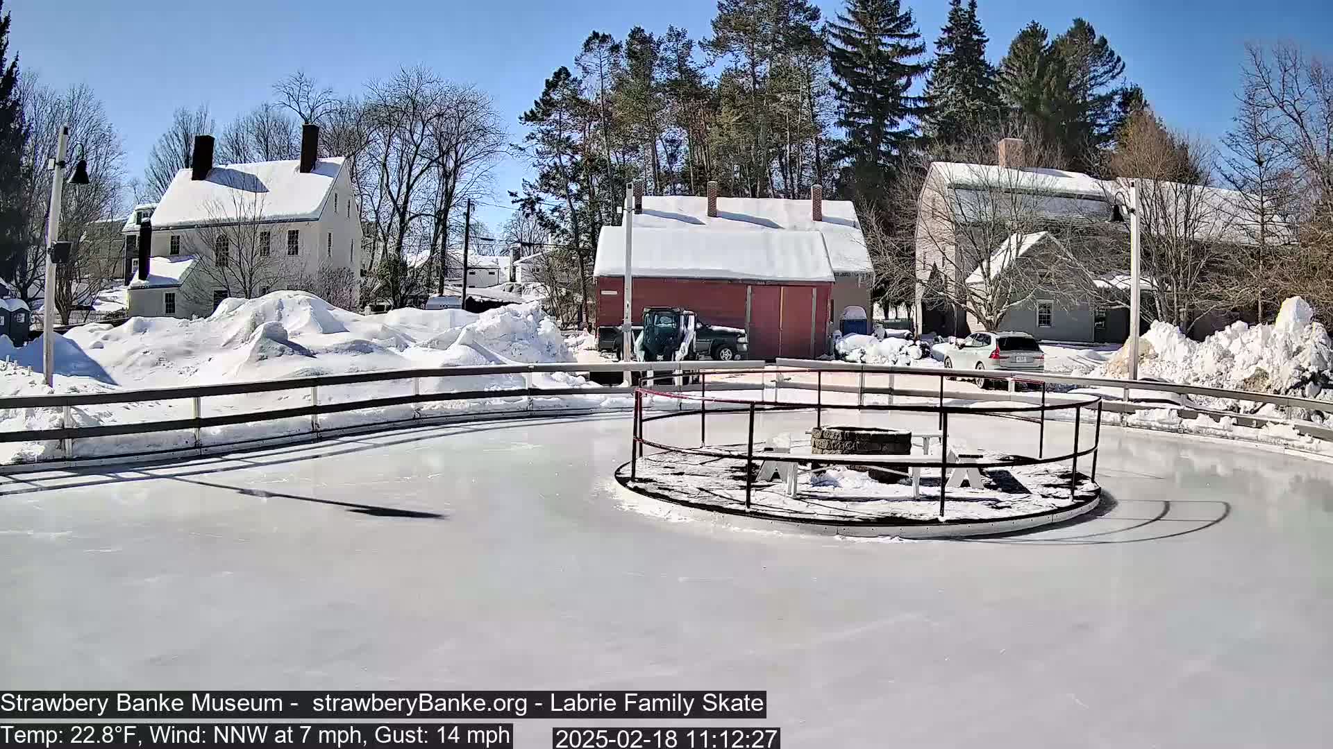 A snow-covered outdoor ice skating rink with several buildings in the background on a sunny day with a light wind.
