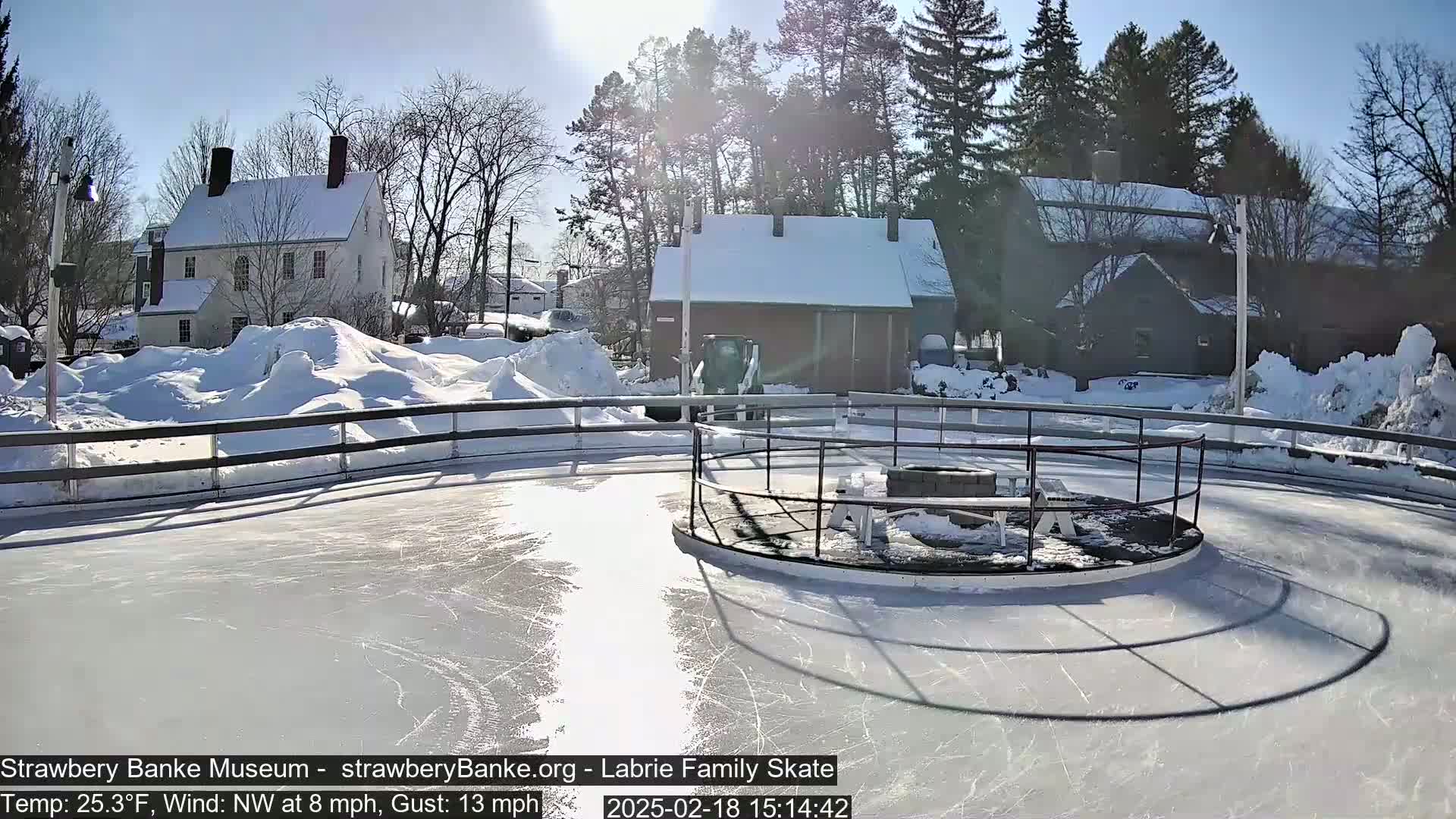 A sunny, snowy day reveals an outdoor ice skating rink surrounded by snow-covered buildings and trees.