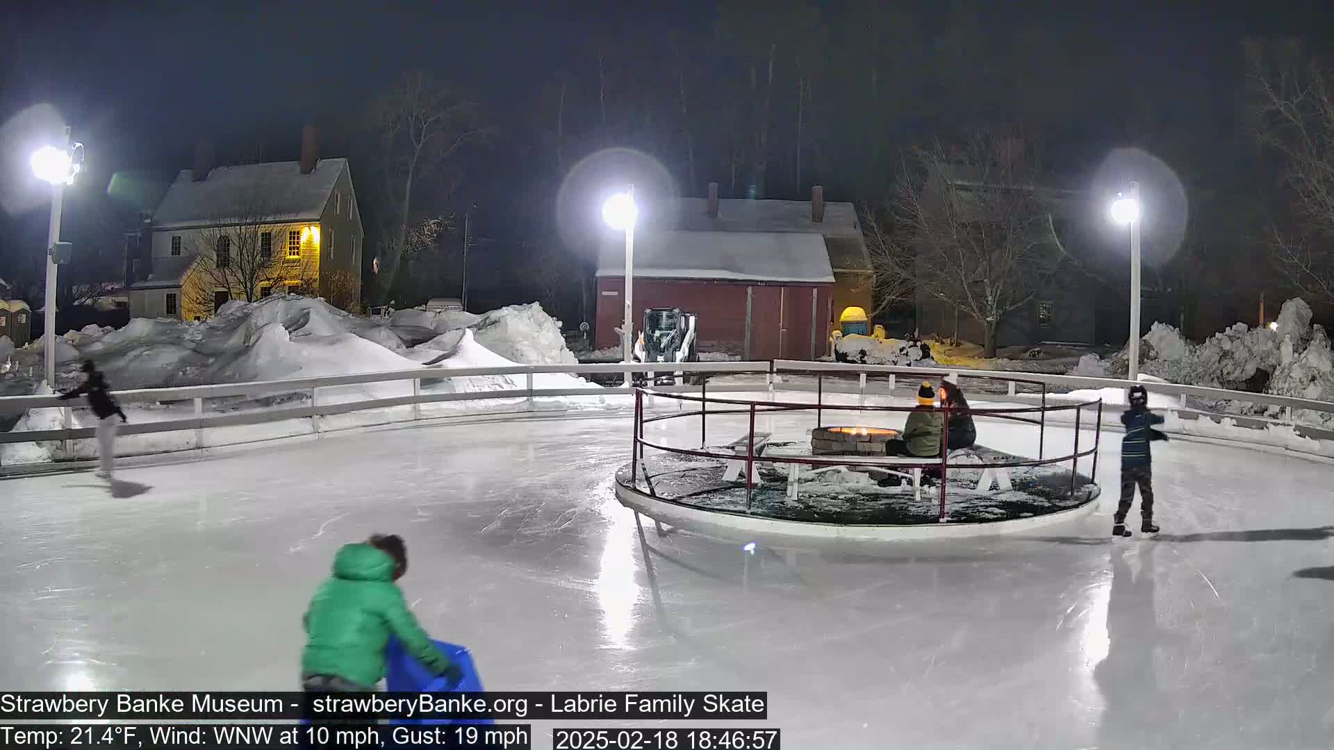 An outdoor ice skating rink at night, illuminated by lights, surrounded by snow and buildings, with several people ice skating and sitting near a fire pit.