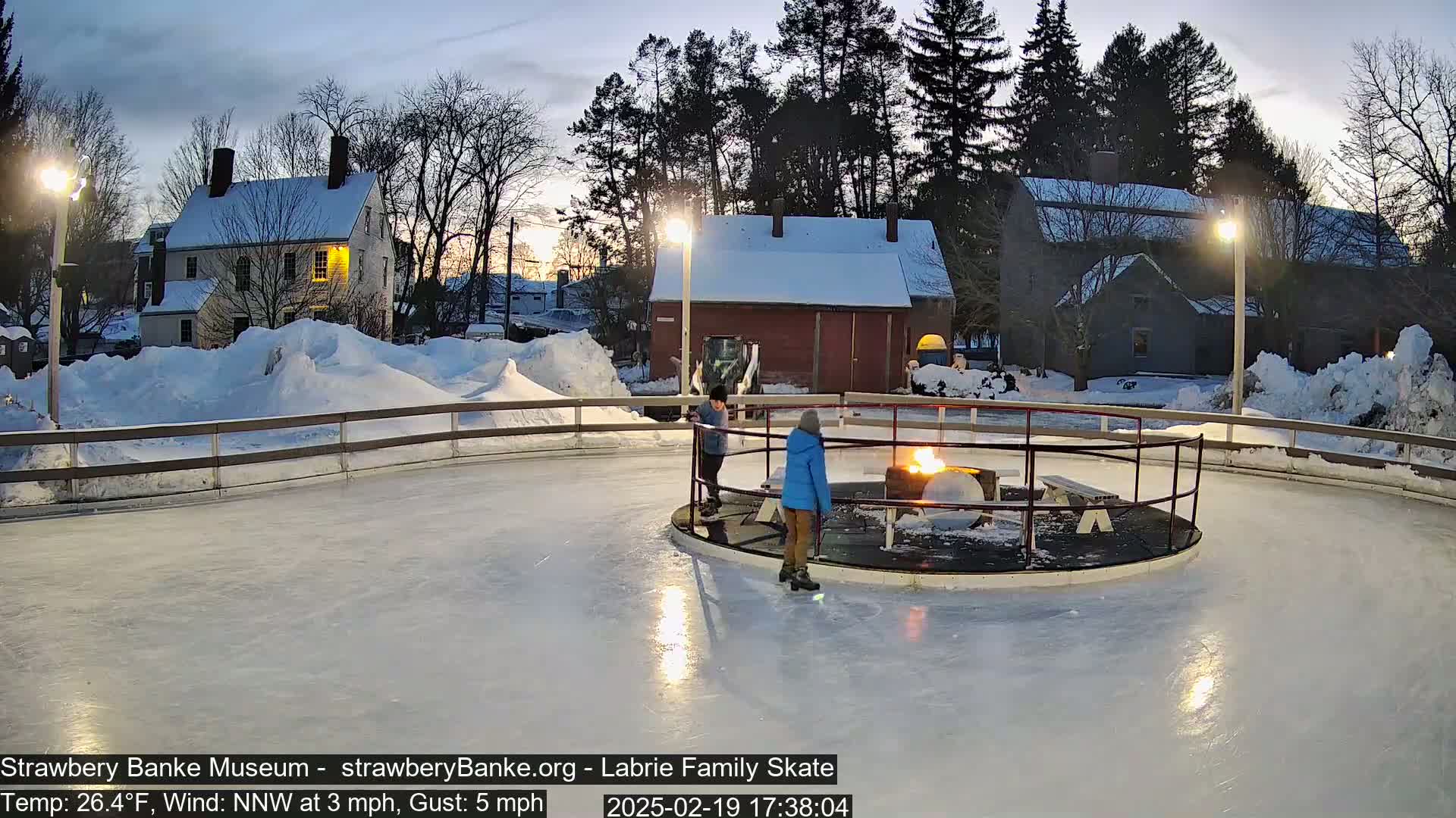 Two people ice skate on a rink at dusk, a fire burning in a central pit, surrounded by snow-covered buildings and trees under a twilight sky.