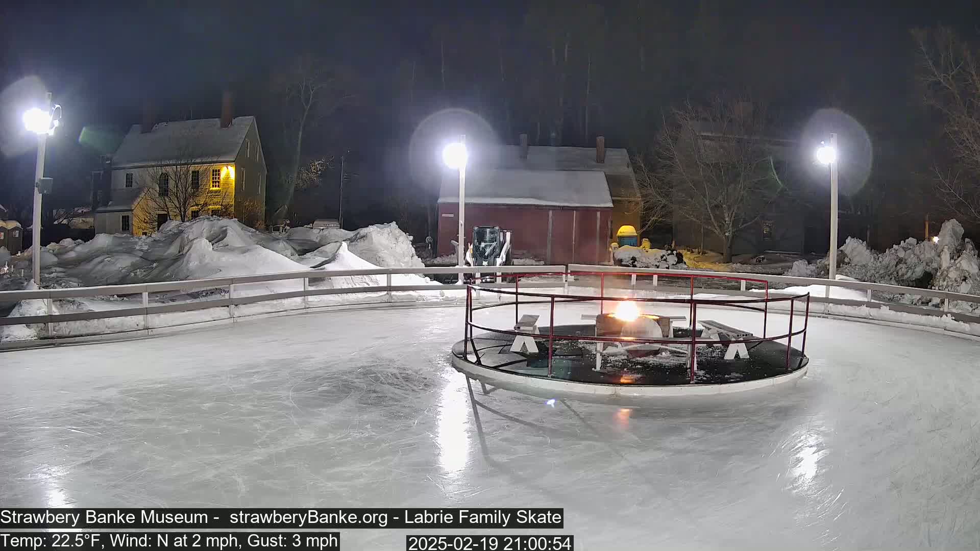 A nighttime view of an outdoor ice skating rink with a fire pit in the center, surrounded by snow-covered buildings and landscaping under lightly clouded skies.