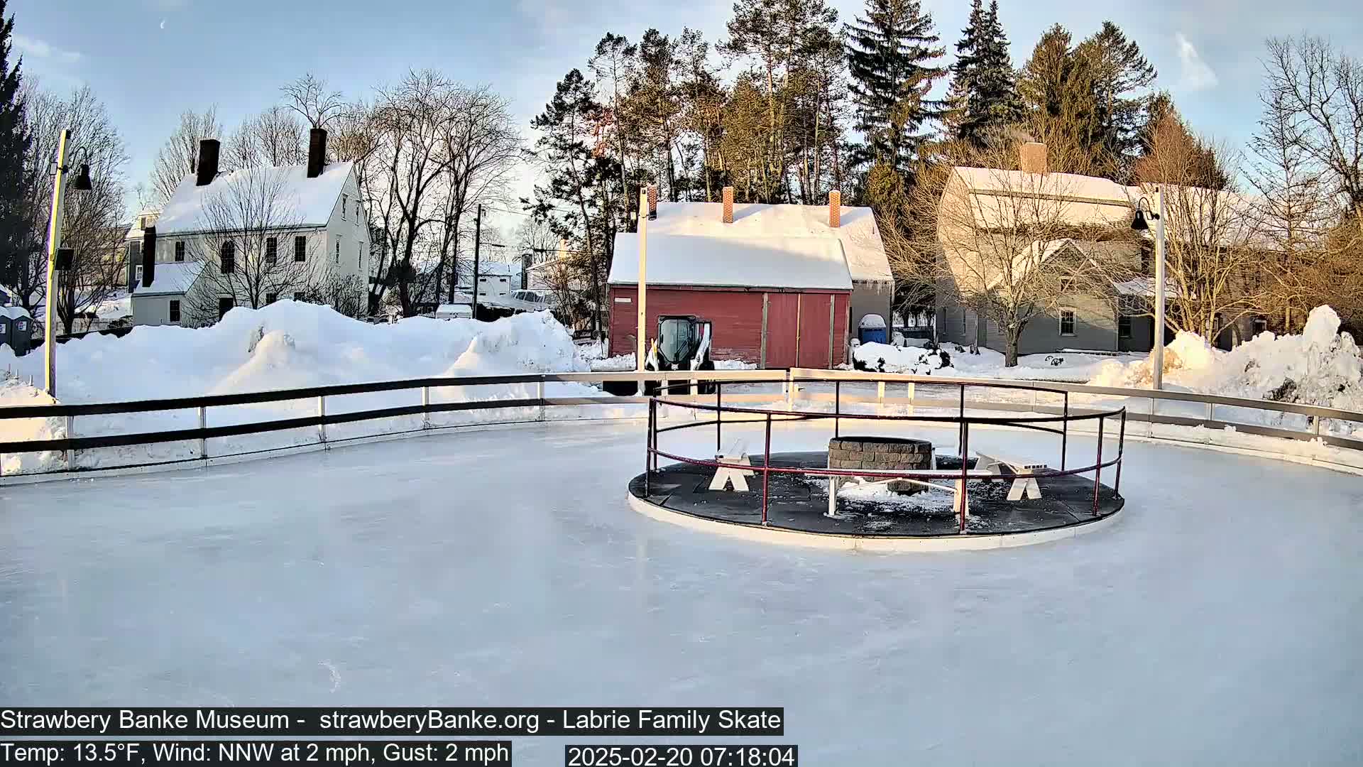 A snow-covered outdoor ice rink, surrounded by snow-covered buildings and trees under a partly sunny sky.