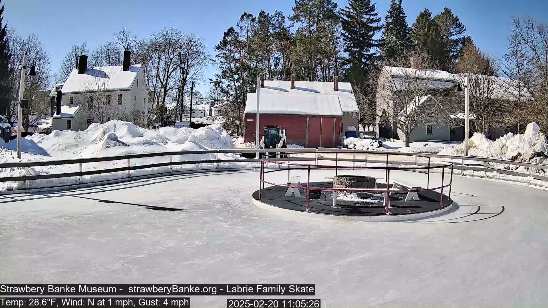 A snow-covered outdoor ice skating rink with a fire pit in the center, surrounded by snow-covered buildings and trees under a sunny sky.
