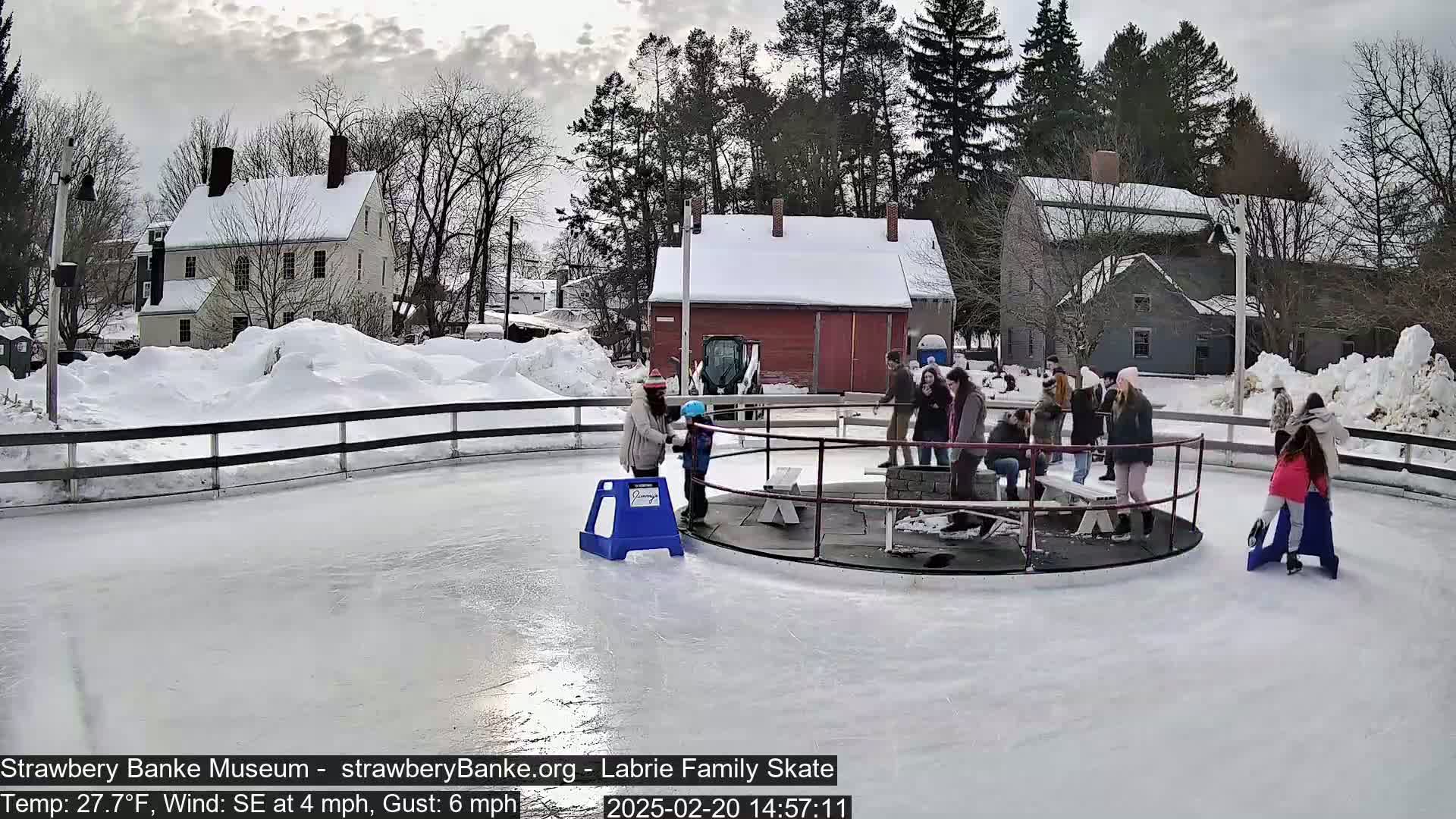People ice skate on an outdoor rink surrounded by snow-covered buildings and trees on a cloudy day with a southeast wind.