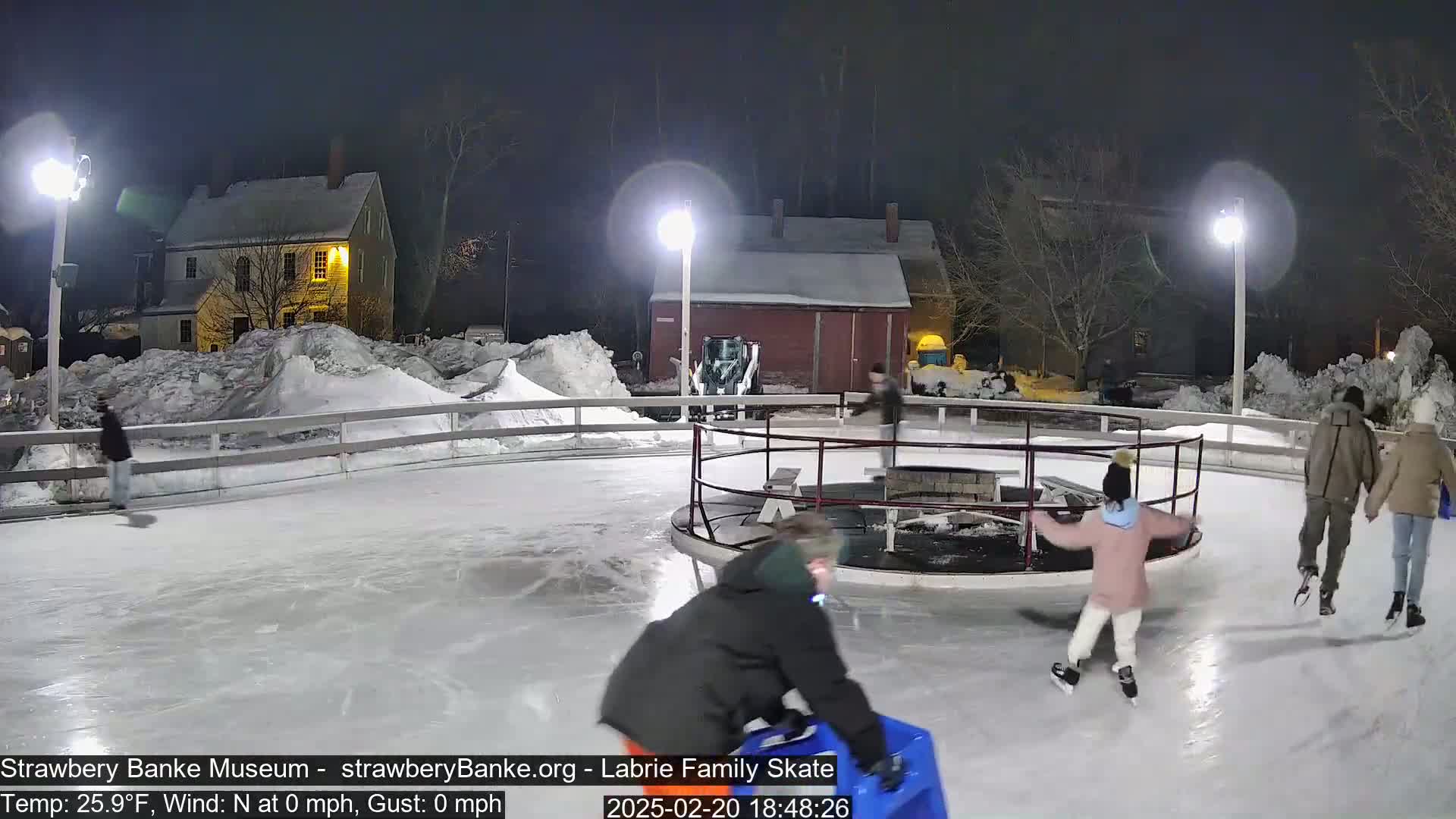 People ice skate on an outdoor rink at night, illuminated by lights, with snow-covered buildings and landscaping in the background.