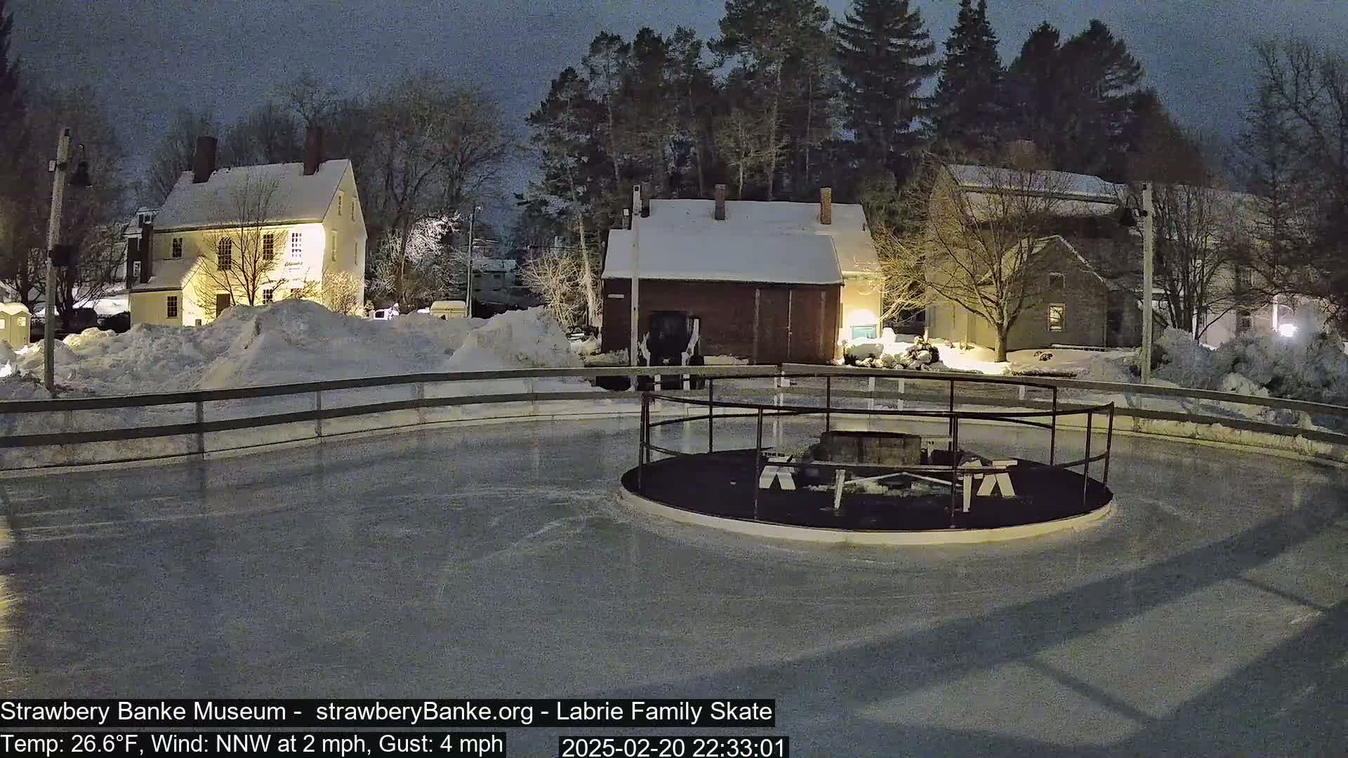 A snow-covered, nighttime scene shows a circular ice skating rink near several illuminated buildings, with a northwest wind at 2 mph and gusts up to 4 mph, and a temperature of 26.6°F.