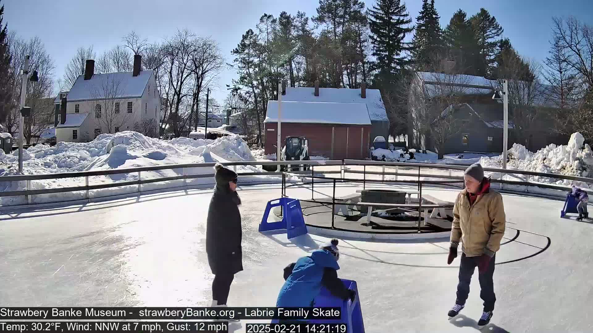 On a sunny, snowy day with a northwest wind, three people ice skate on an outdoor rink near snow-covered buildings.