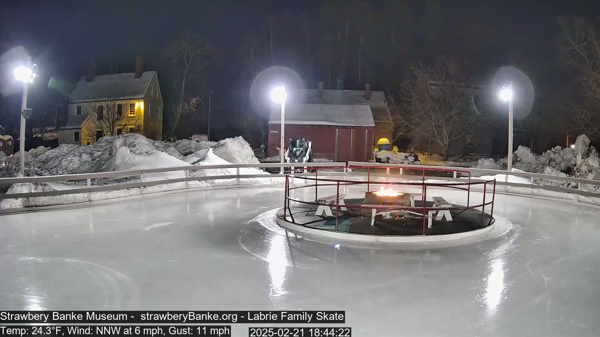 A nighttime view of an outdoor ice skating rink with a fire pit in the center, surrounded by snow and illuminated by several lights.