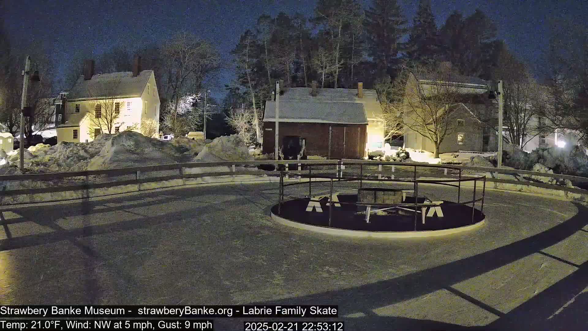 A snow-covered, nighttime scene shows a fenced-in circular skating rink near several illuminated colonial-style buildings under a dark, mostly clear sky, with a northwest wind at 5 mph and gusts up to 9 mph.