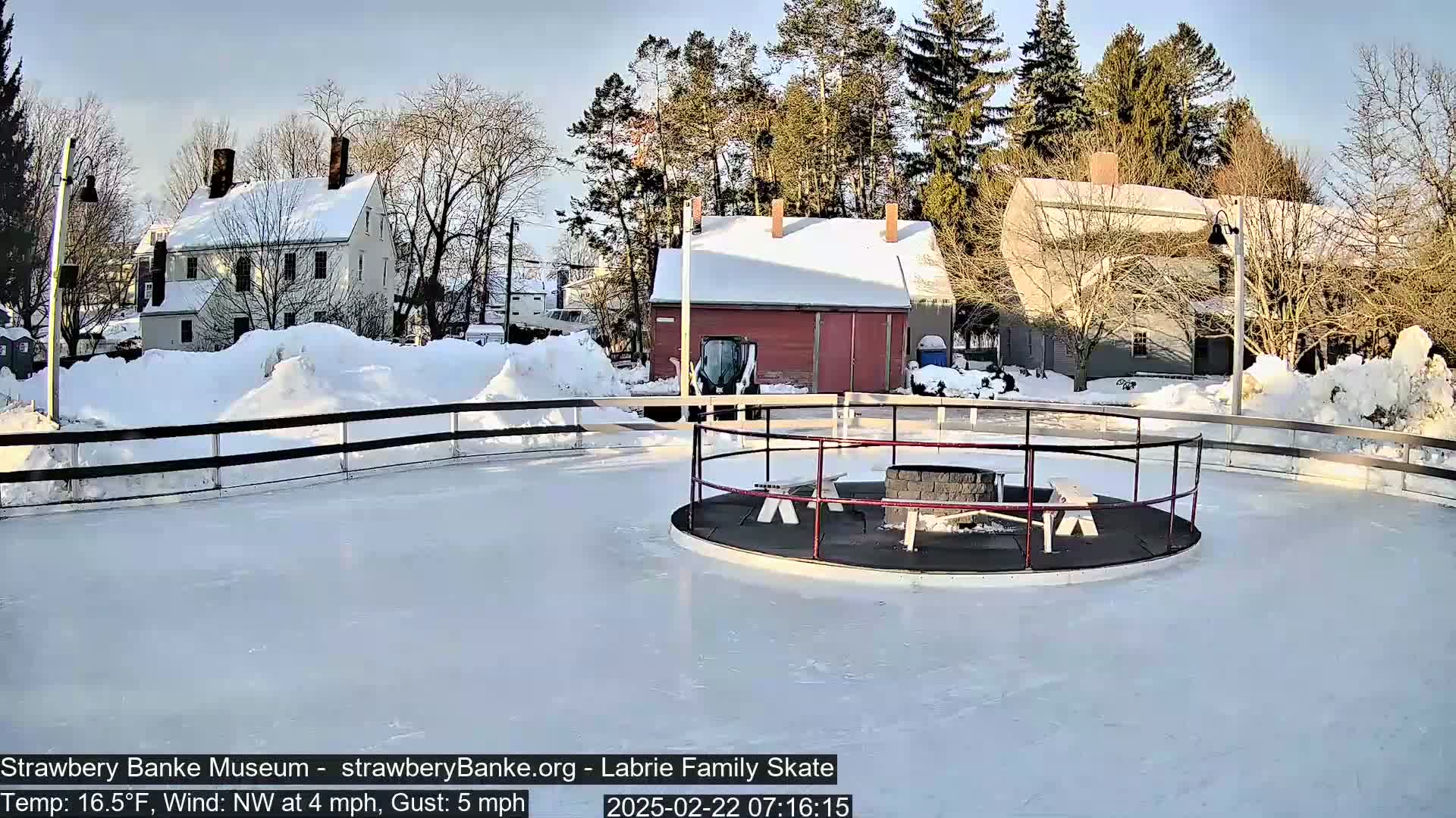 A snowy outdoor ice rink, surrounded by snow-covered buildings and trees, under a sunny sky with a northwest wind.