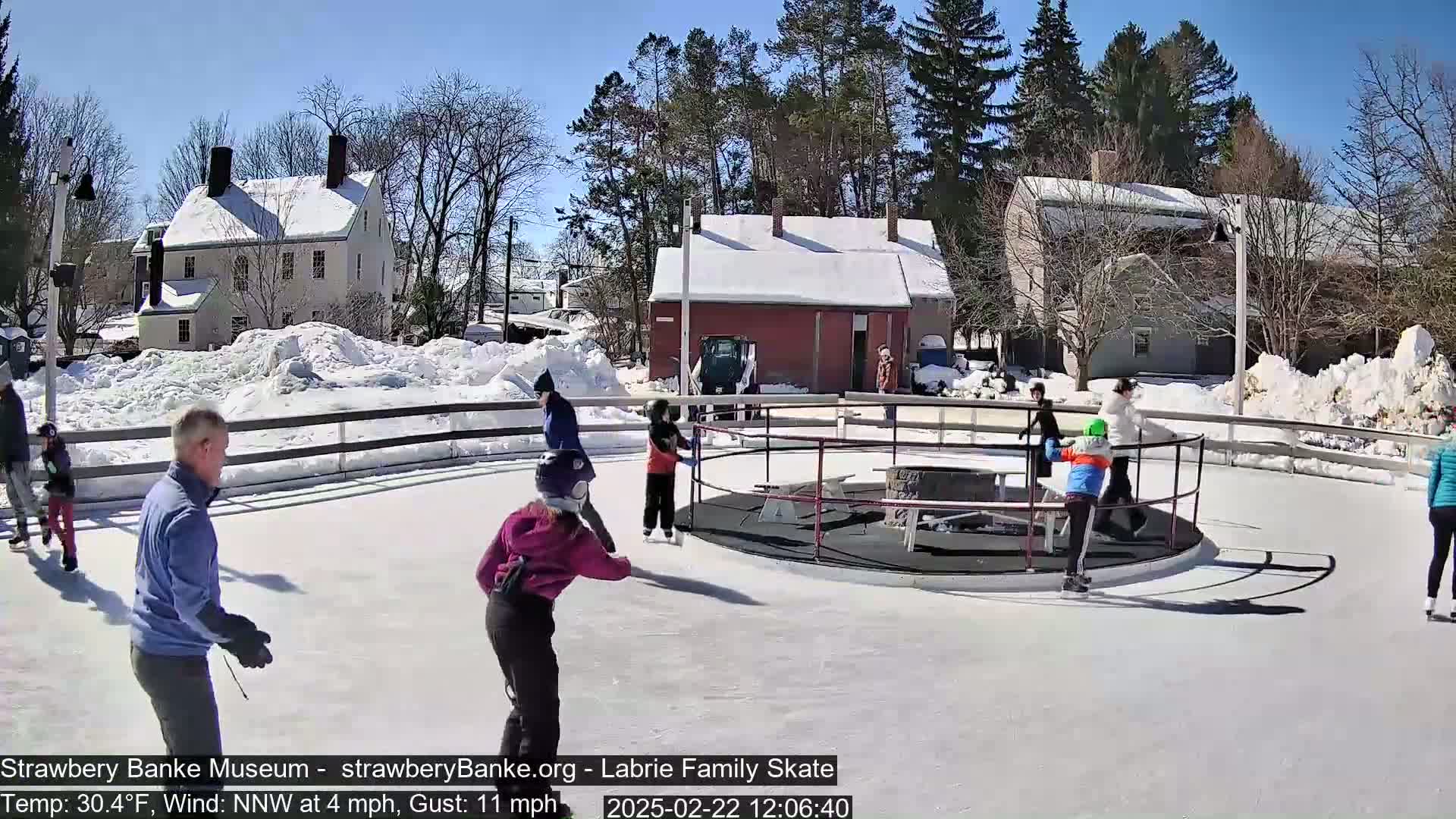 A sunny day shows several people ice skating on an outdoor rink, surrounded by snow-covered buildings and trees.