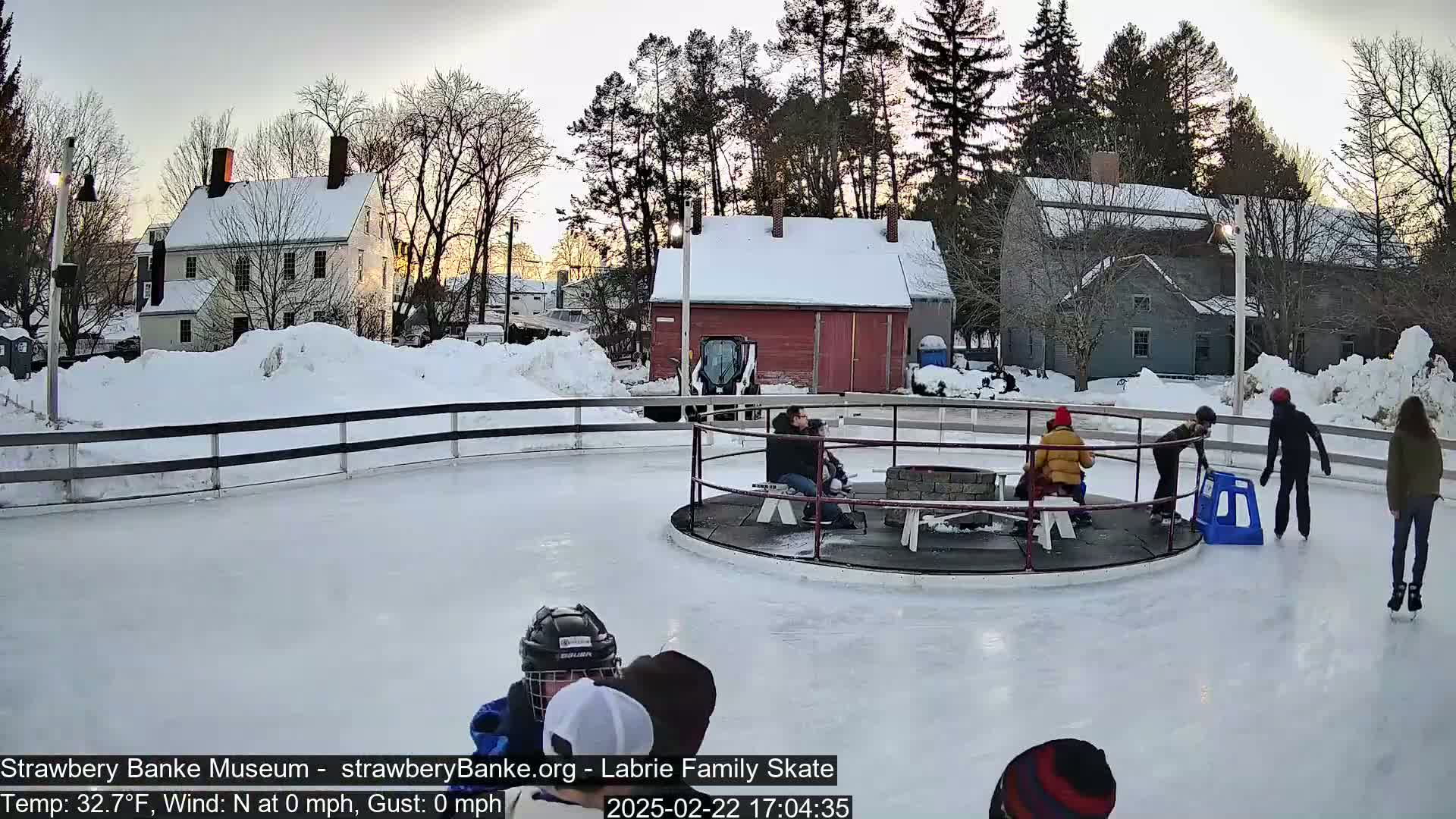 An outdoor ice skating rink in a snowy setting, surrounded by snow-covered buildings and trees under a twilight sky.