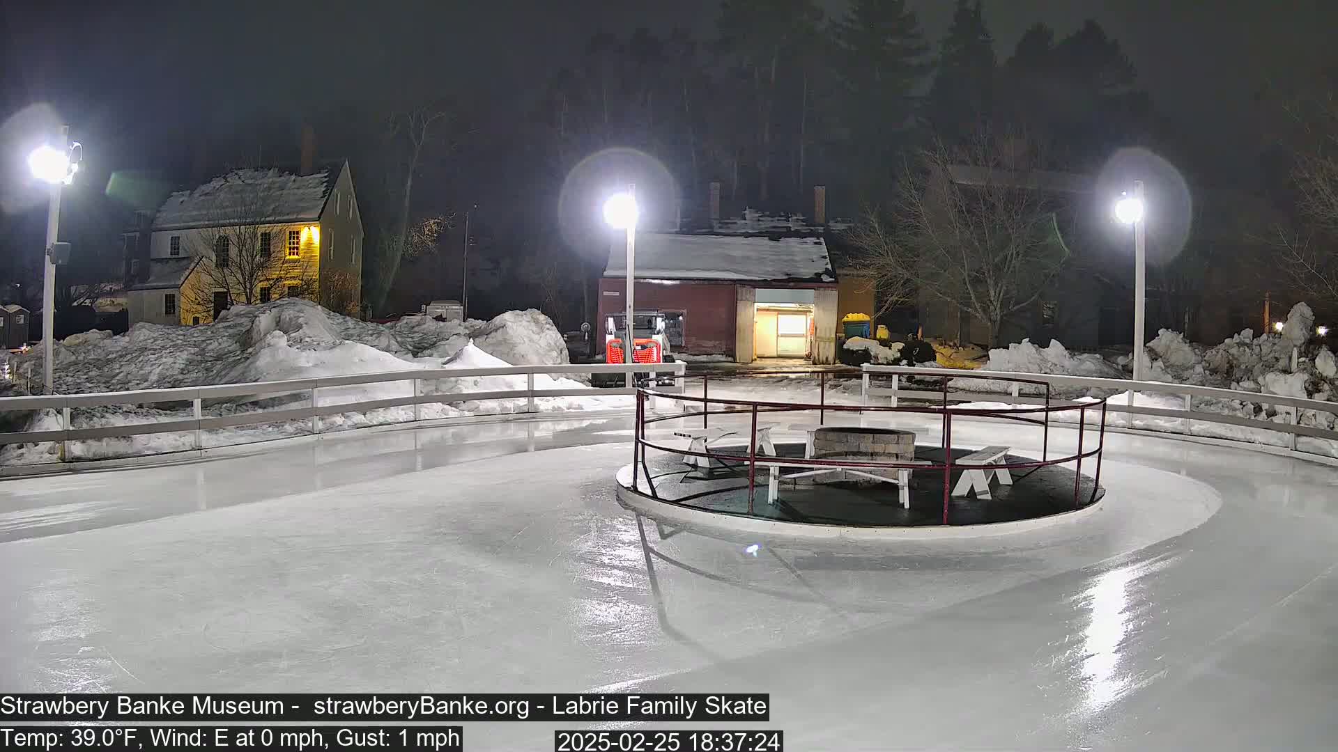 An outdoor ice skating rink illuminated at night, surrounded by snow and buildings, with a small fire pit in the center.