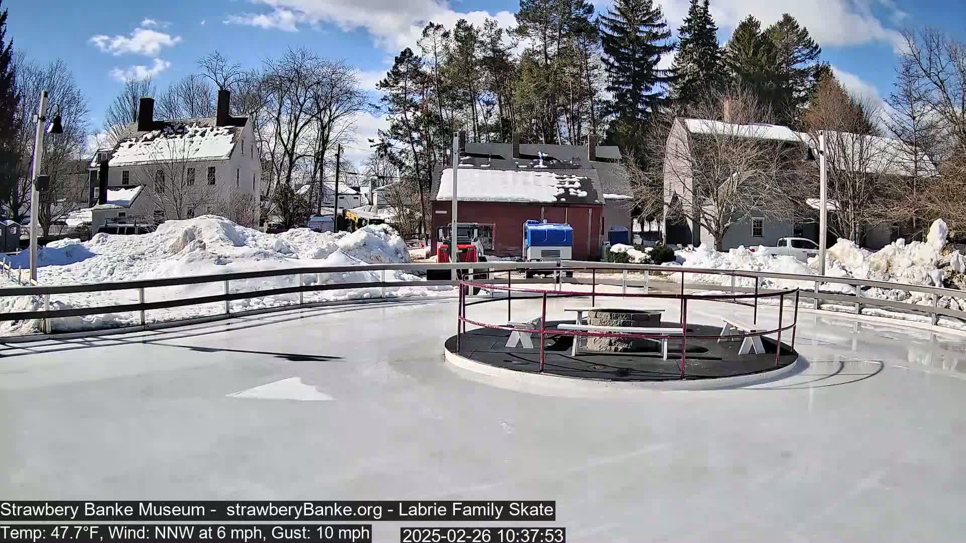 A snow-covered outdoor ice skating rink, surrounded by buildings and trees under a partly sunny sky.