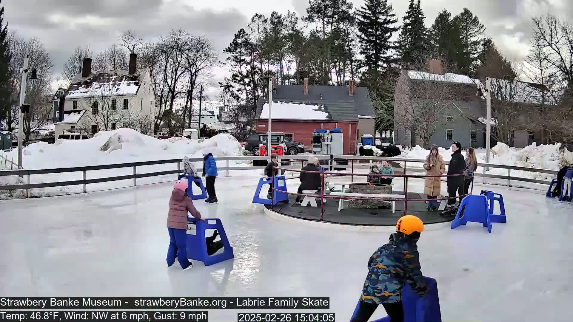 People ice skate on an outdoor rink surrounded by snow and bare trees under a cloudy sky.