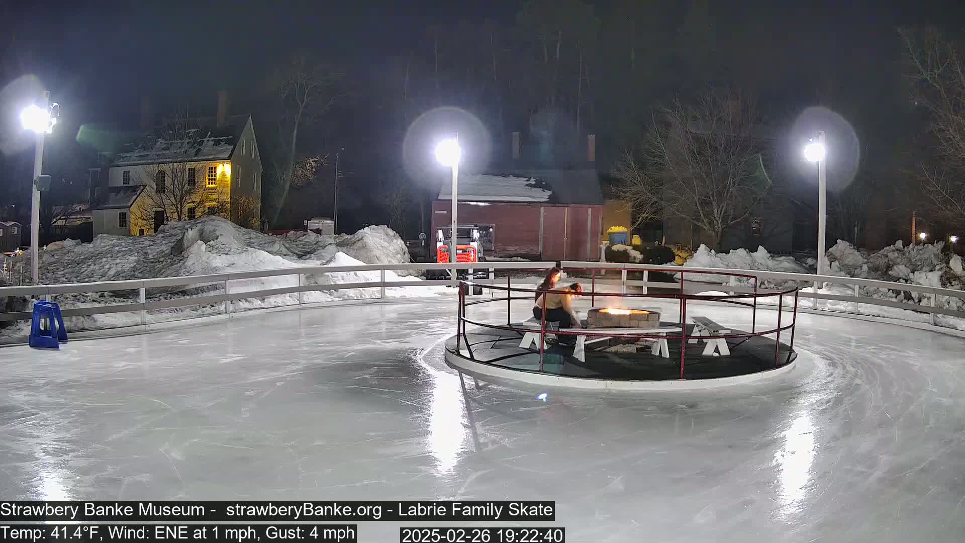 An outdoor ice skating rink at night, lit by several lamps, with a fire pit in the center and two people sitting nearby, surrounded by snow.