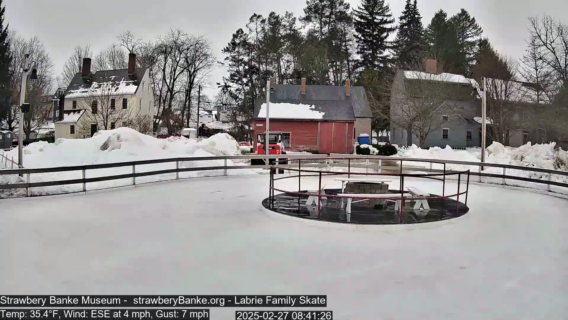 A snow-covered outdoor skating rink with a fire pit and picnic tables surrounded by snow-dusted buildings and trees under an overcast sky.