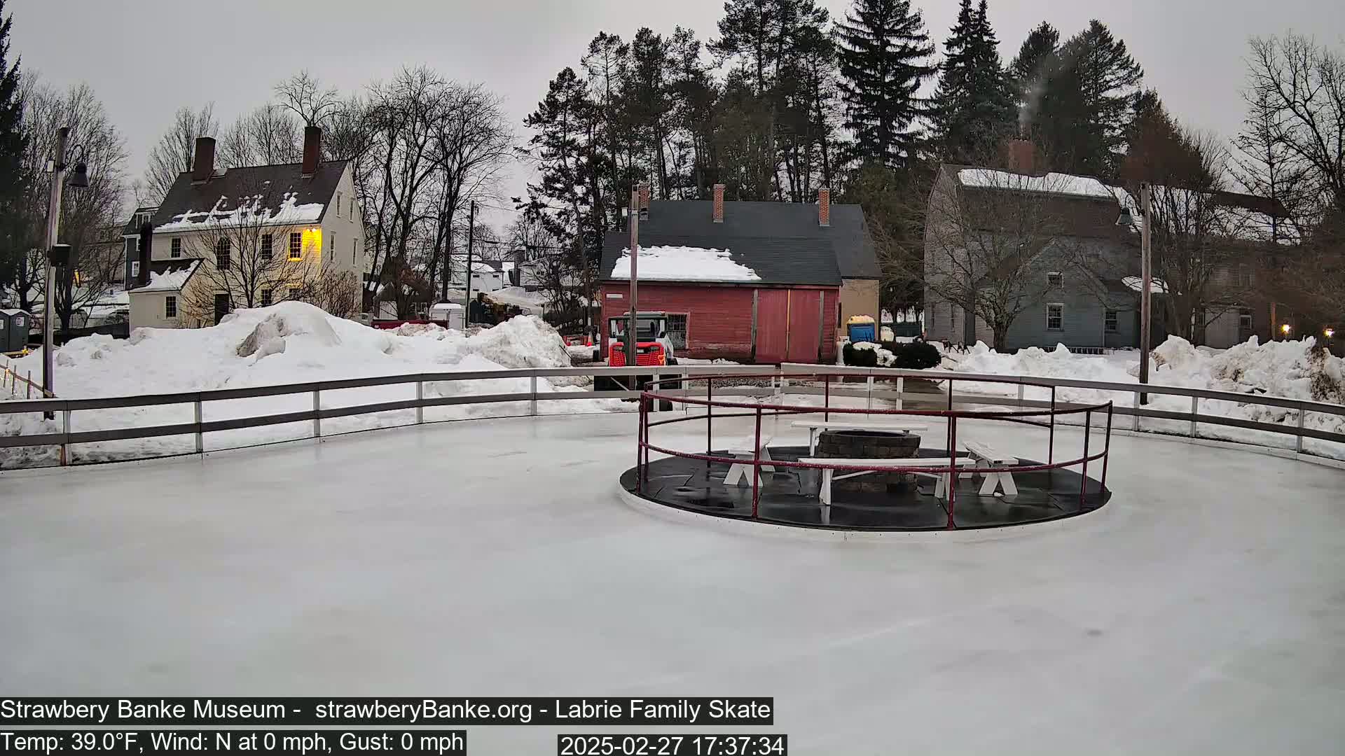 A snow-covered outdoor ice rink, encircled by a fence, features a fire pit and picnic tables in the center, with several buildings and snowdrifts visible in the background under a cloudy sky.